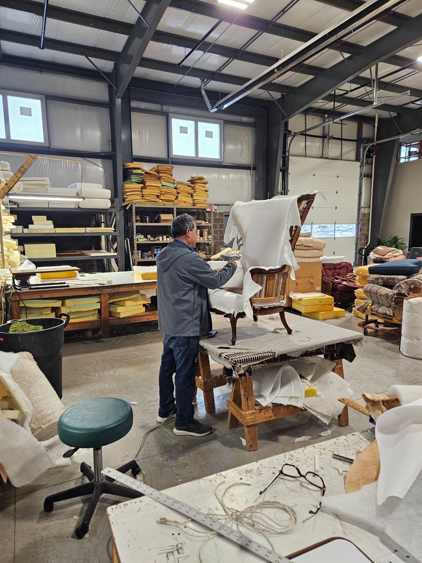 Man reupholstering a chair in a workshop. He's wearing a jacket and working at a table, with supplies and other chairs nearby.