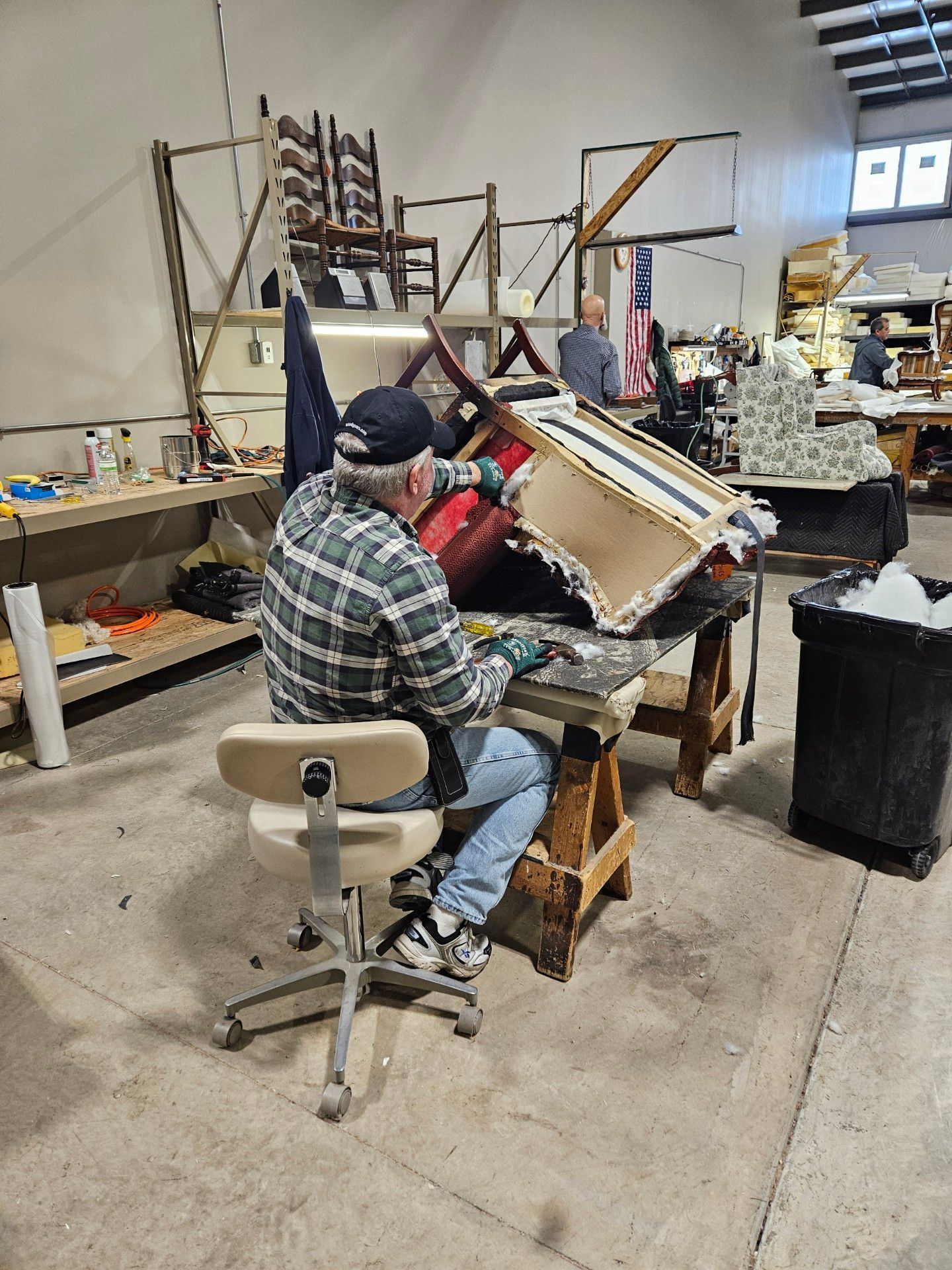 A person reupholsters furniture in a workshop. They wear a plaid shirt, jeans, and a cap, working on a chair frame.