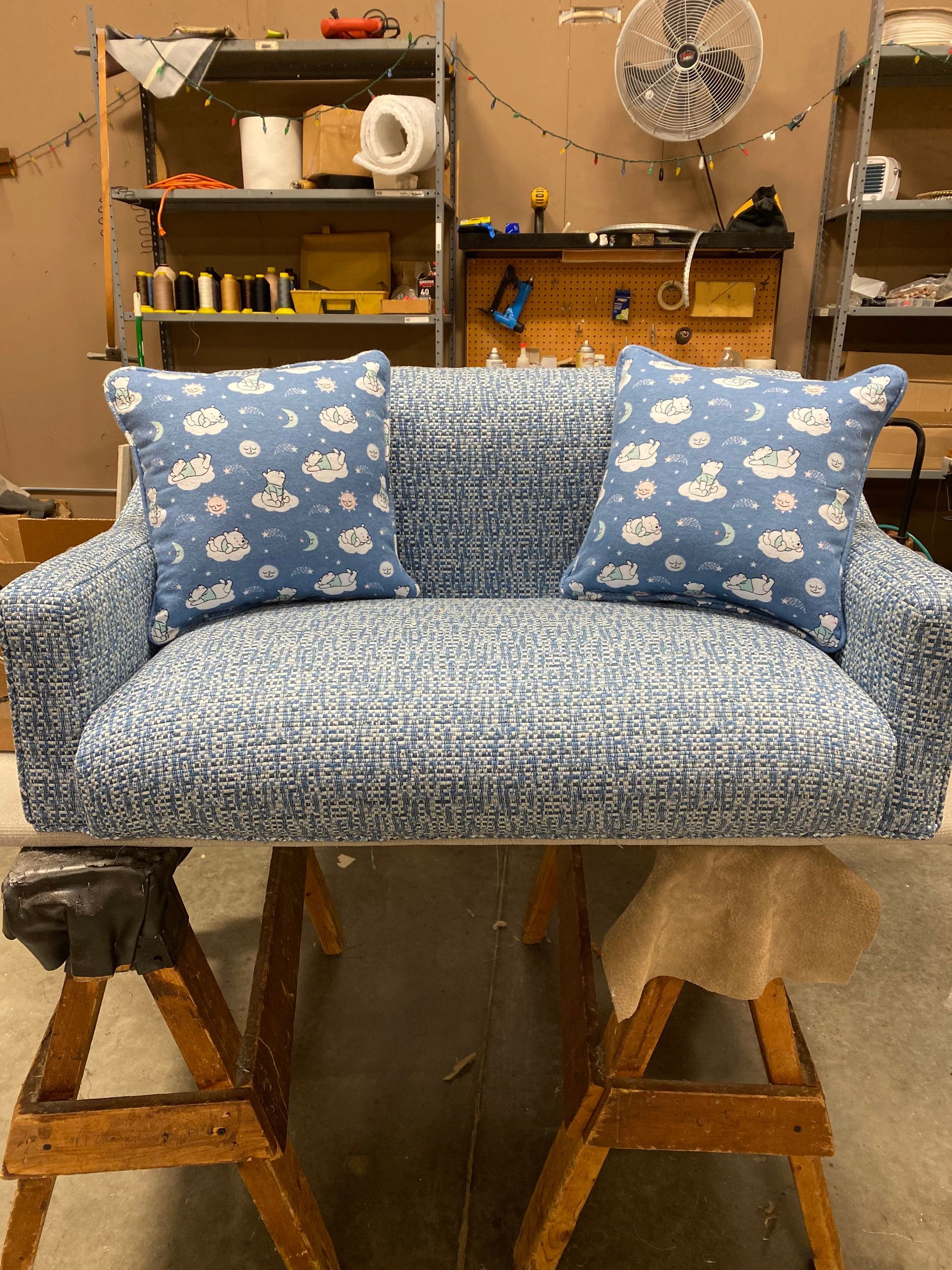 A newly upholstered blue couch with two patterned pillows, set on wooden sawhorses in a workshop.