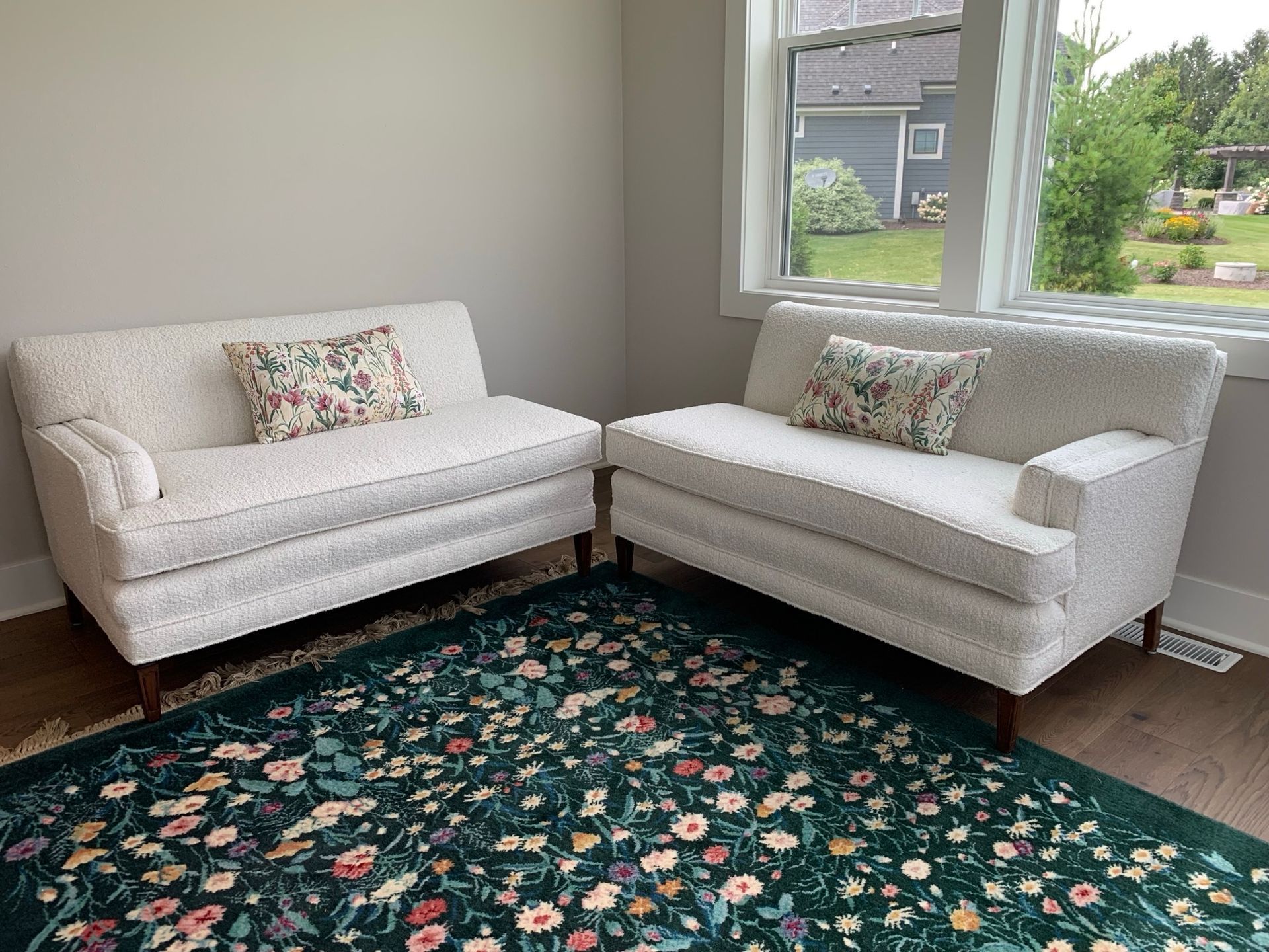 Two off white loveseats with floral pillows sit on a floral rug, near a window with a view.