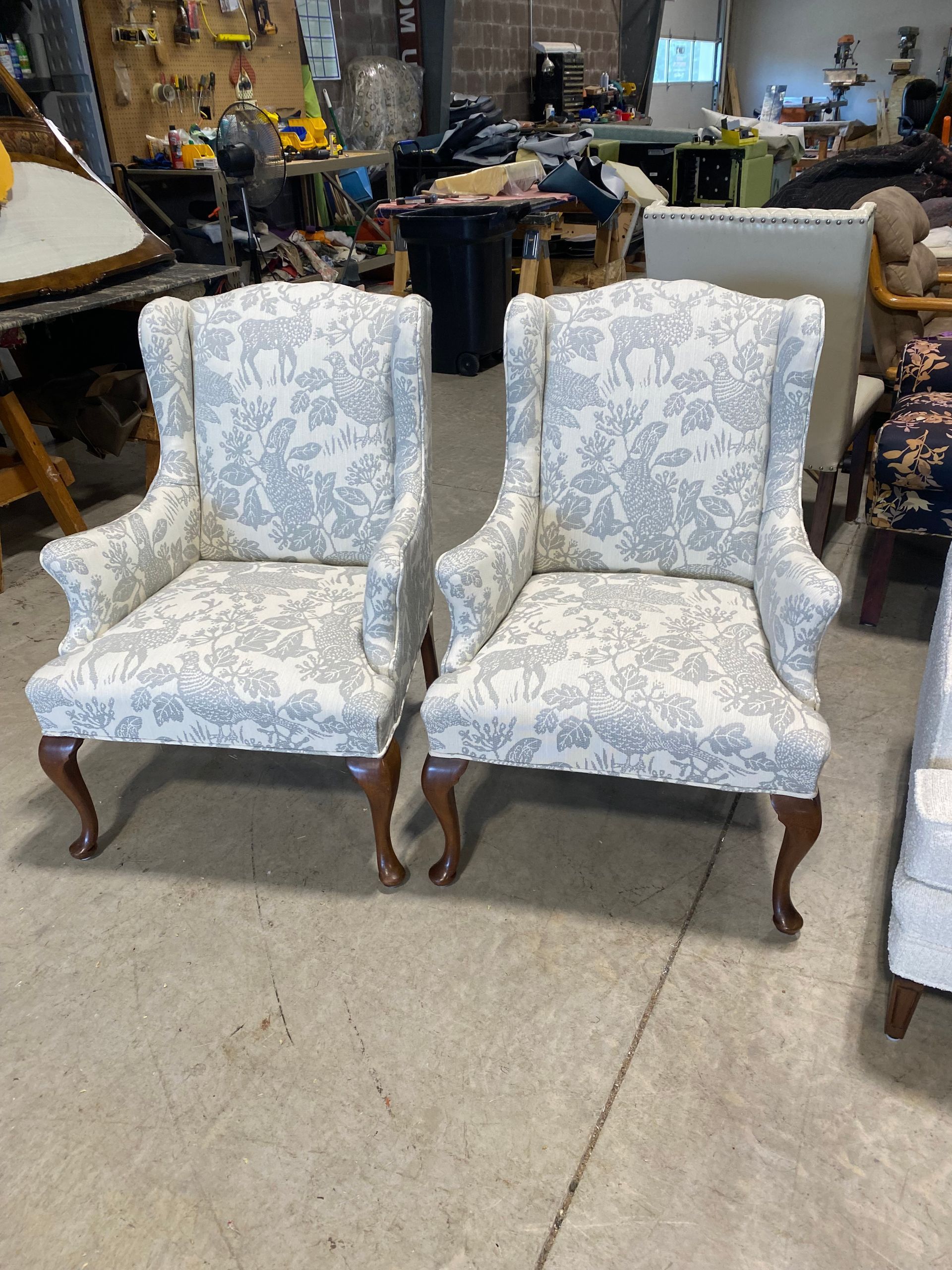Two wingback chairs with blue floral print fabric, brown legs, on a concrete floor.