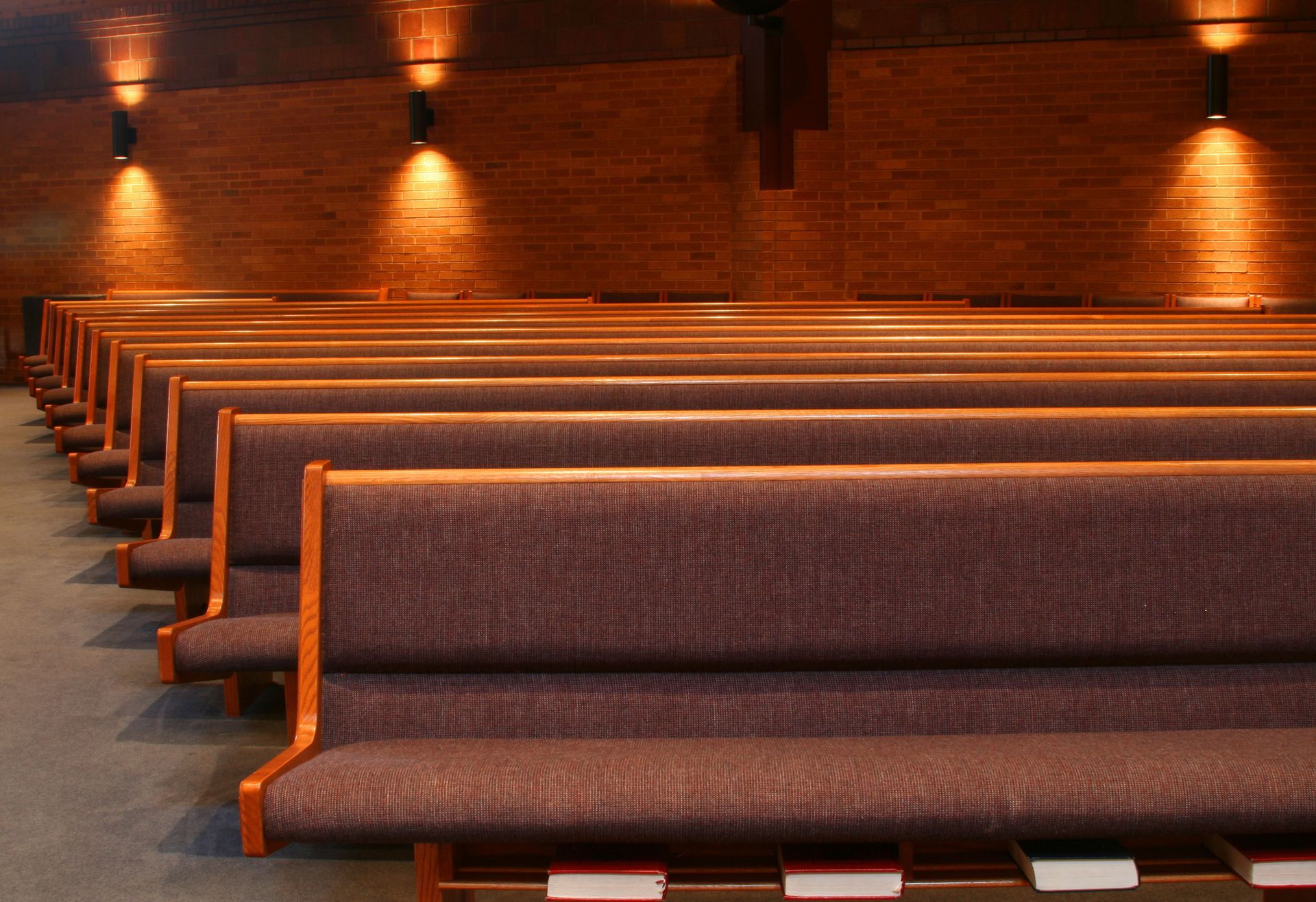 Rows of empty church pews facing a brick wall lit by overhead lights. Wooden trim and brown upholstery.