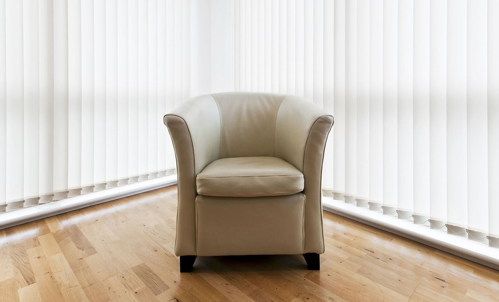 Cream-colored armchair in a corner of a room with light-colored vertical blinds and hardwood floor.