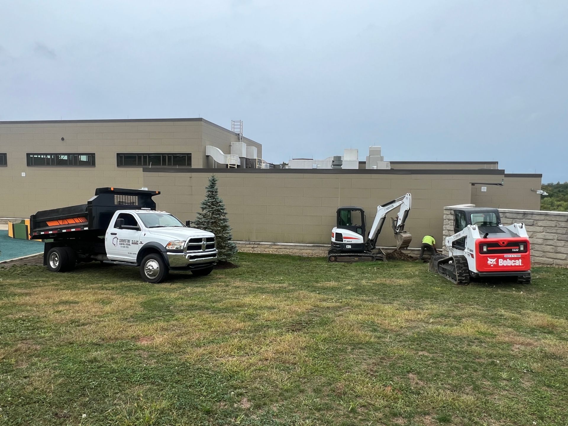 A dump truck and an excavator are parked in a grassy field in front of a building.