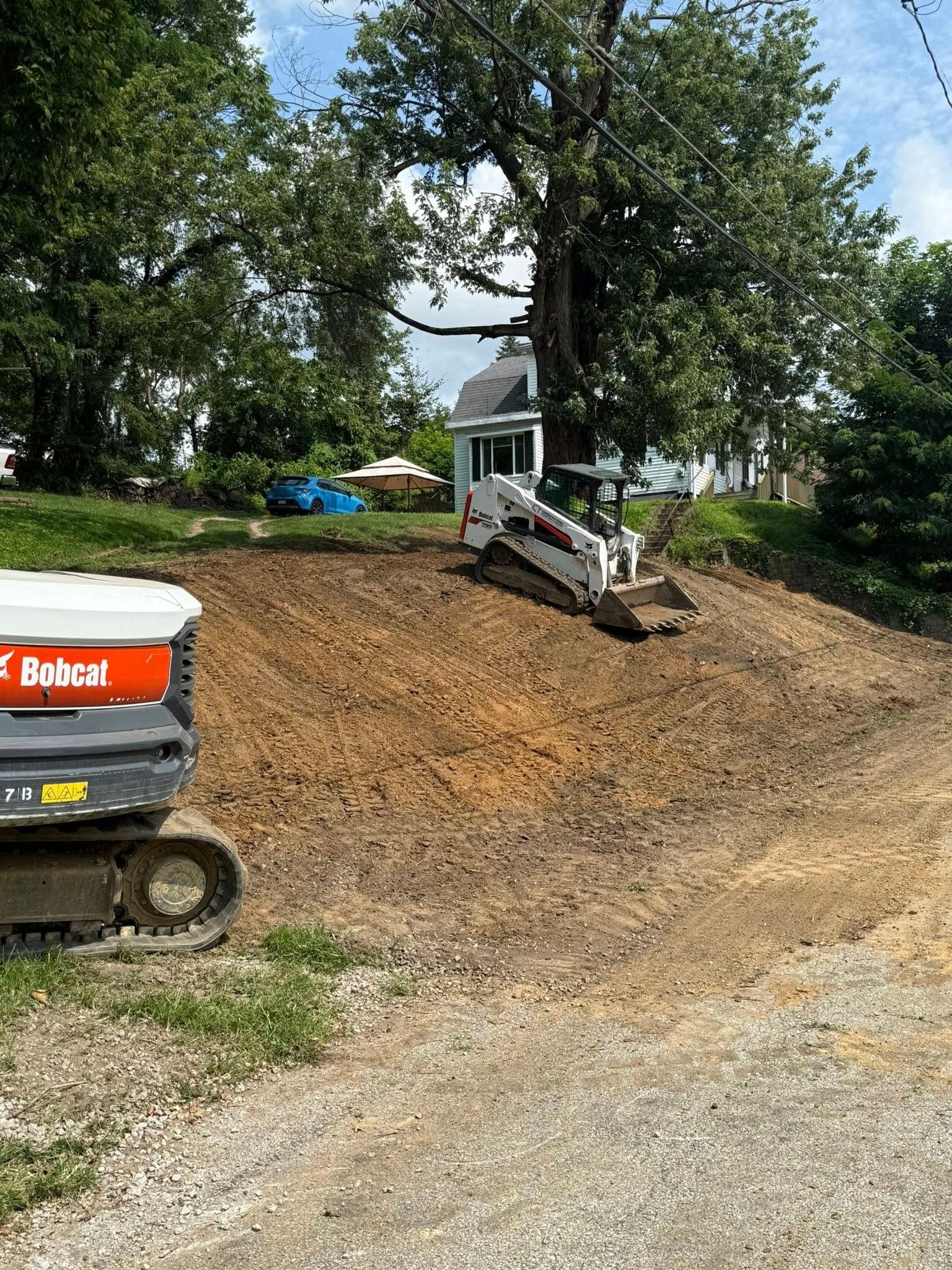 Two Bobcat construction machines on a dirt mound. Trees and a light blue house are in the background on a sunny day.