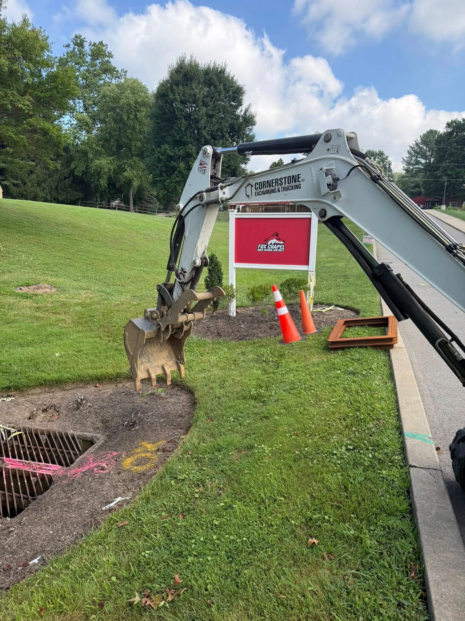 An excavator digs in a grassy area beside a sidewalk, near a sign and traffic cones.