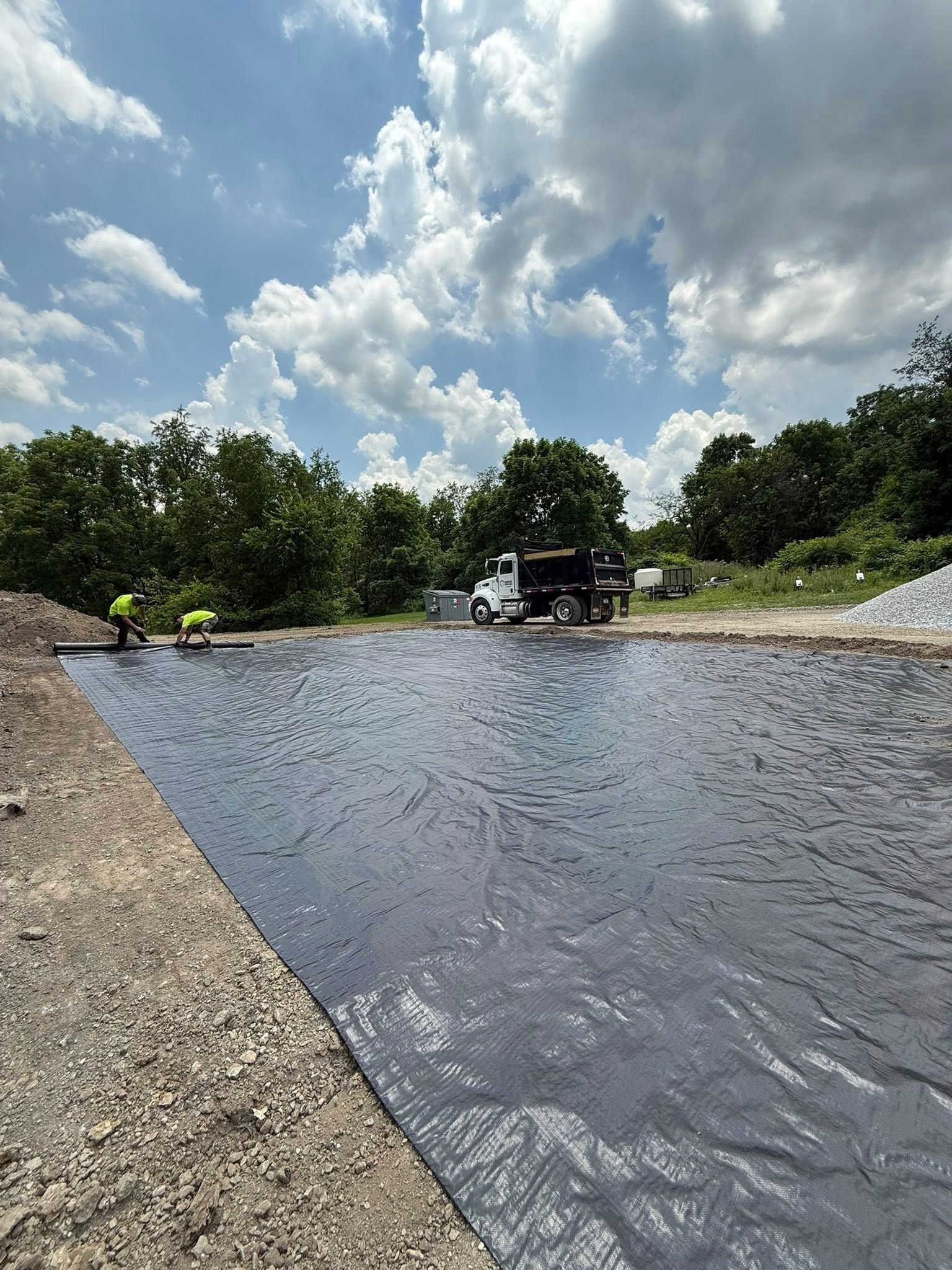 Workers laying black fabric in a construction site on a sunny day. A dump truck sits nearby.