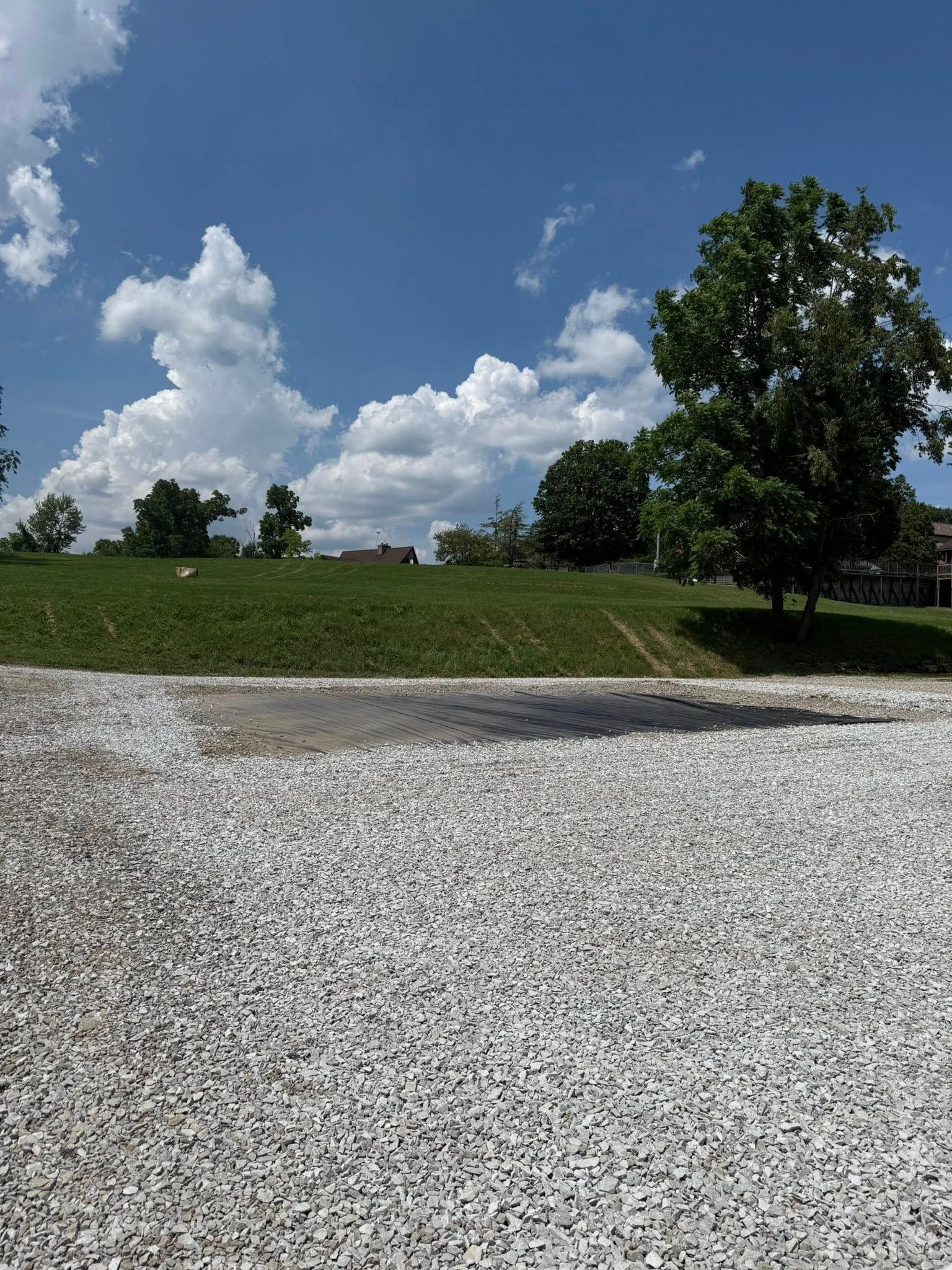 White gravel foreground leads to a grassy hill with trees under a blue sky with fluffy white clouds.
