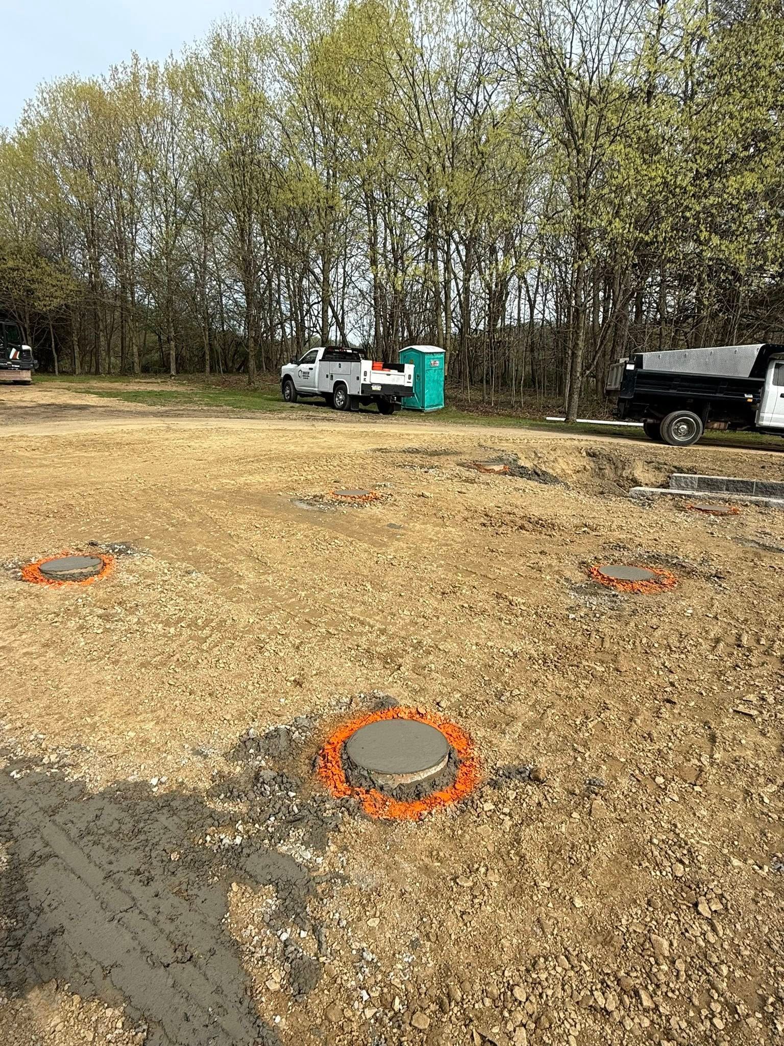 Construction site with a concrete lid surrounded by orange markers. Two white trucks and a portable toilet are visible.