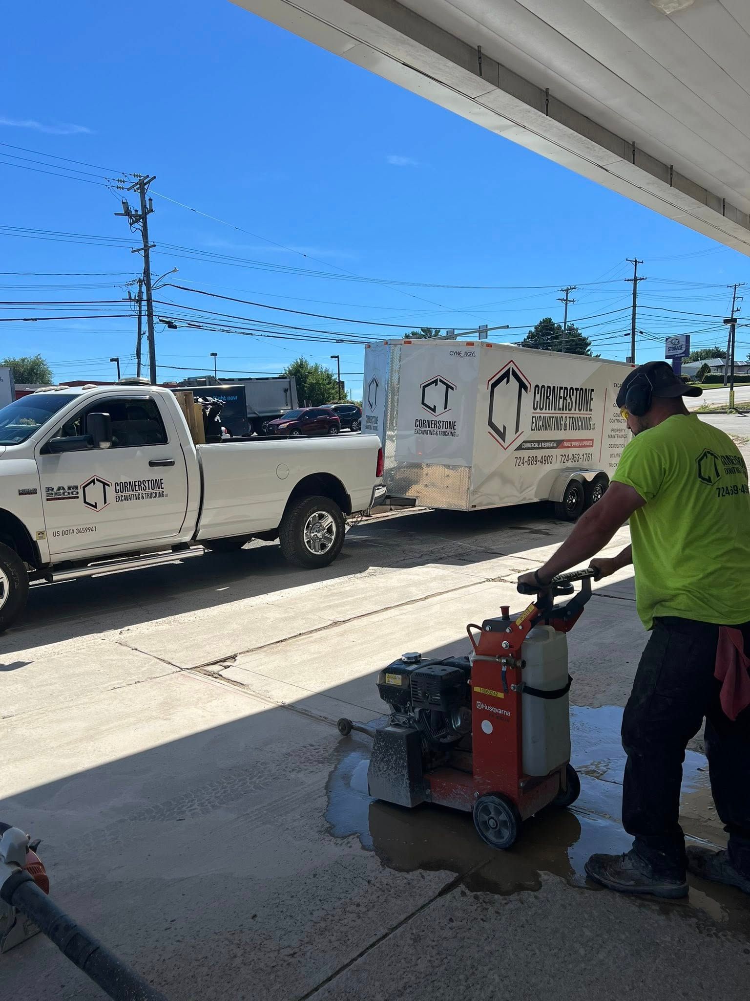 A worker cuts concrete with a machine at a gas station, a truck and trailer are parked nearby on a sunny day.