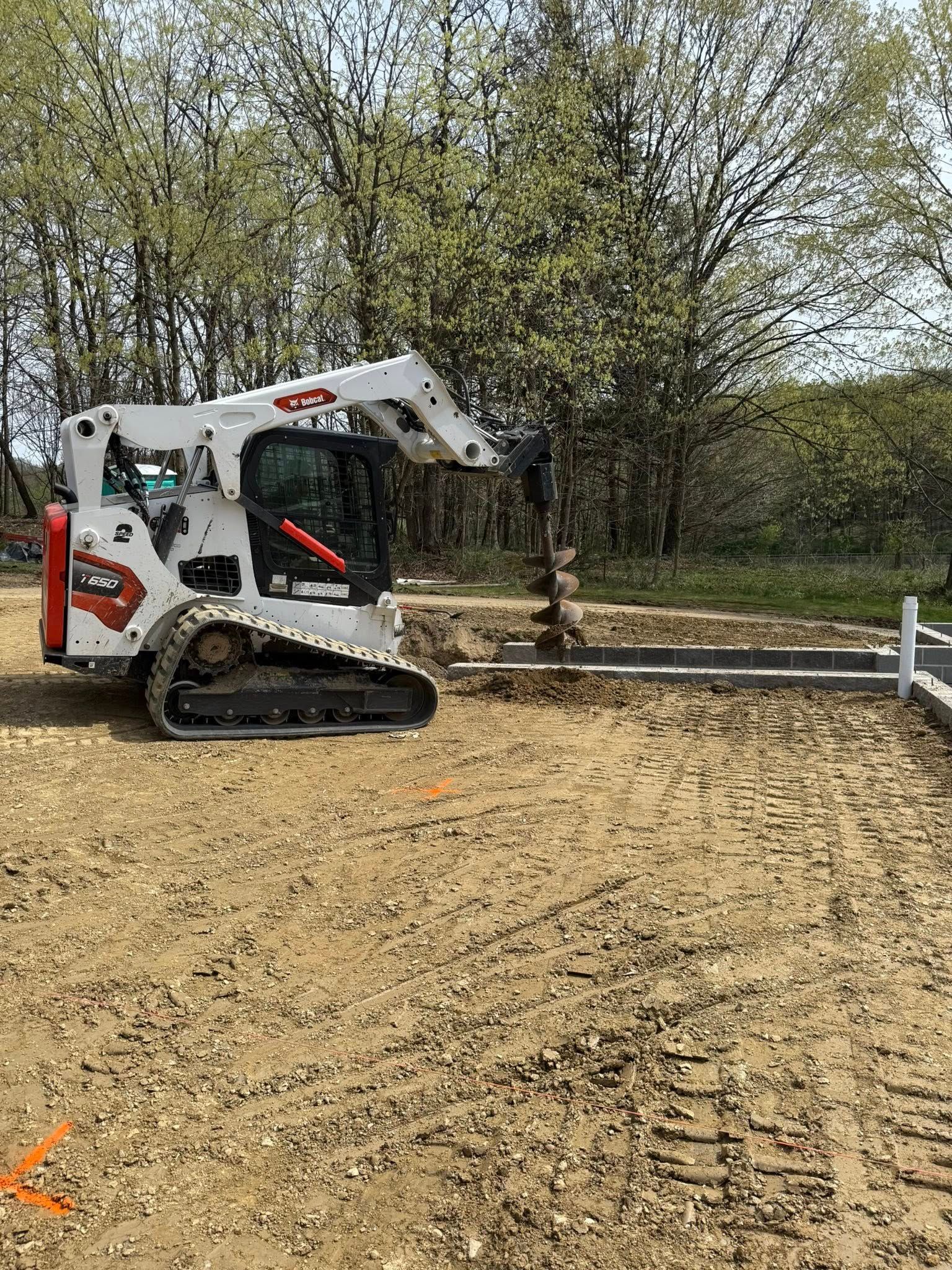 A white Bobcat skid-steer loader on a construction site, next to a trench. The ground is brown, and trees are in the background.