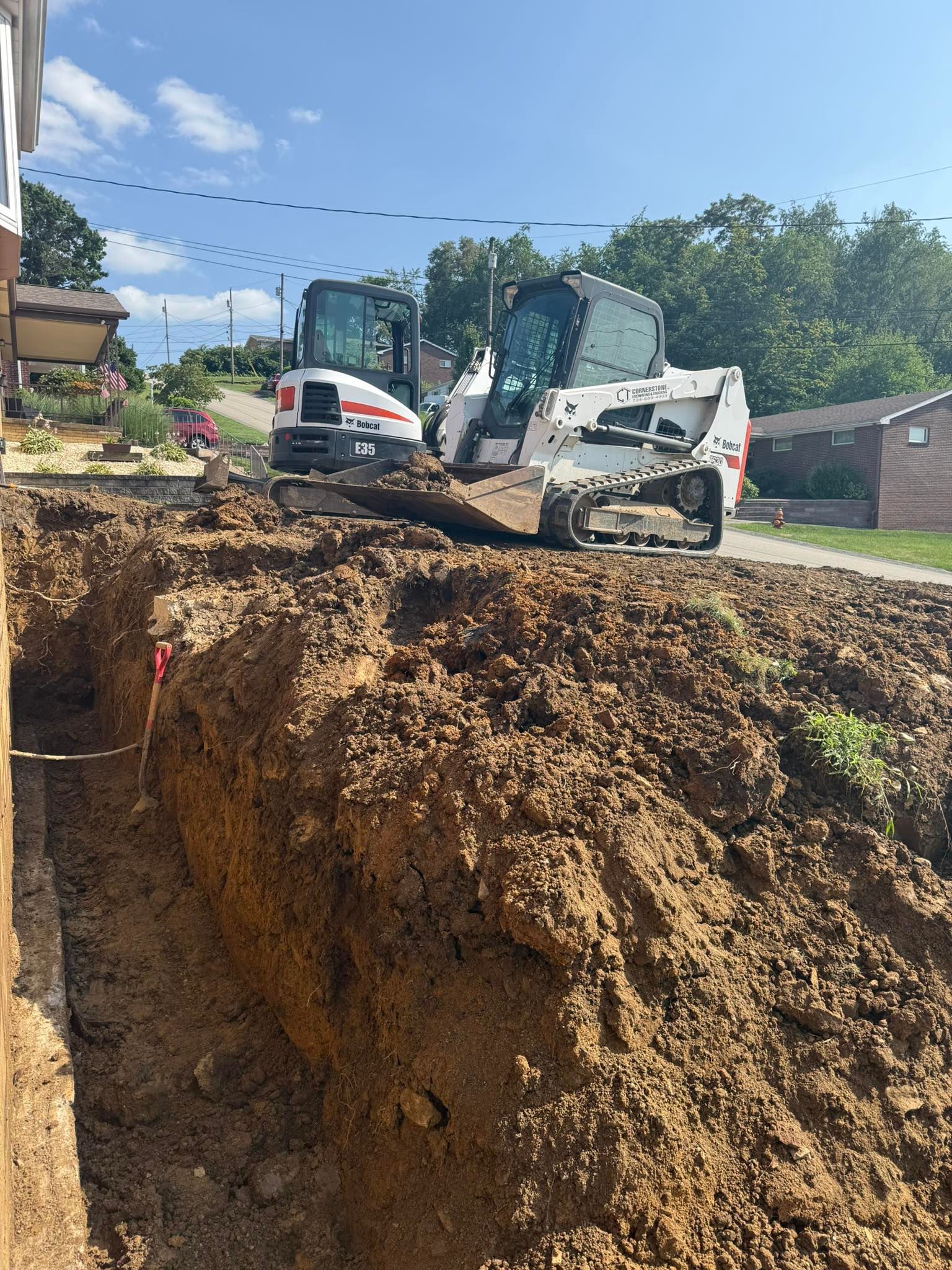 Two Bobcat construction vehicles excavating a trench next to a building on a sunny day. A large pile of dirt is in the foreground.