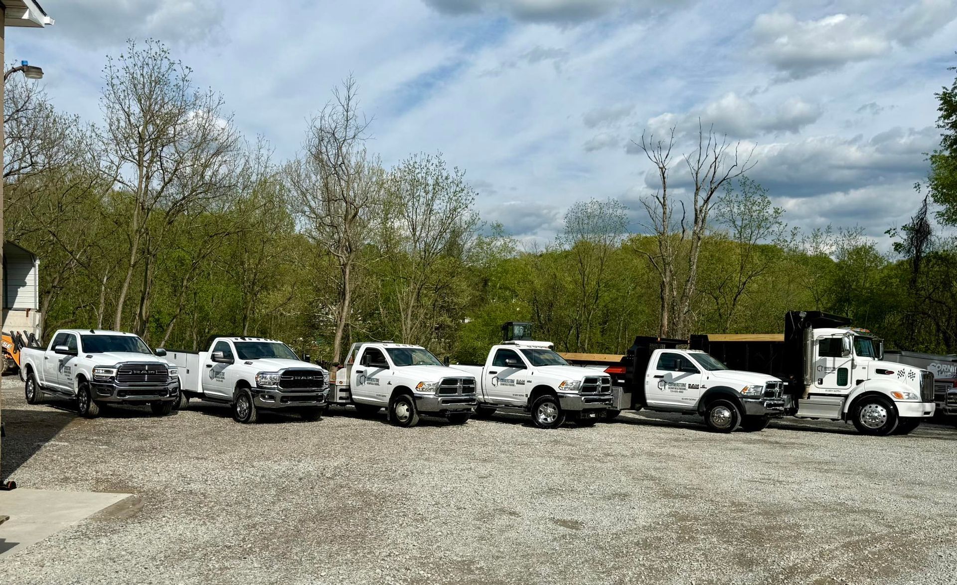 Lineup of six white work trucks parked on gravel with a backdrop of trees under a partly cloudy sky.