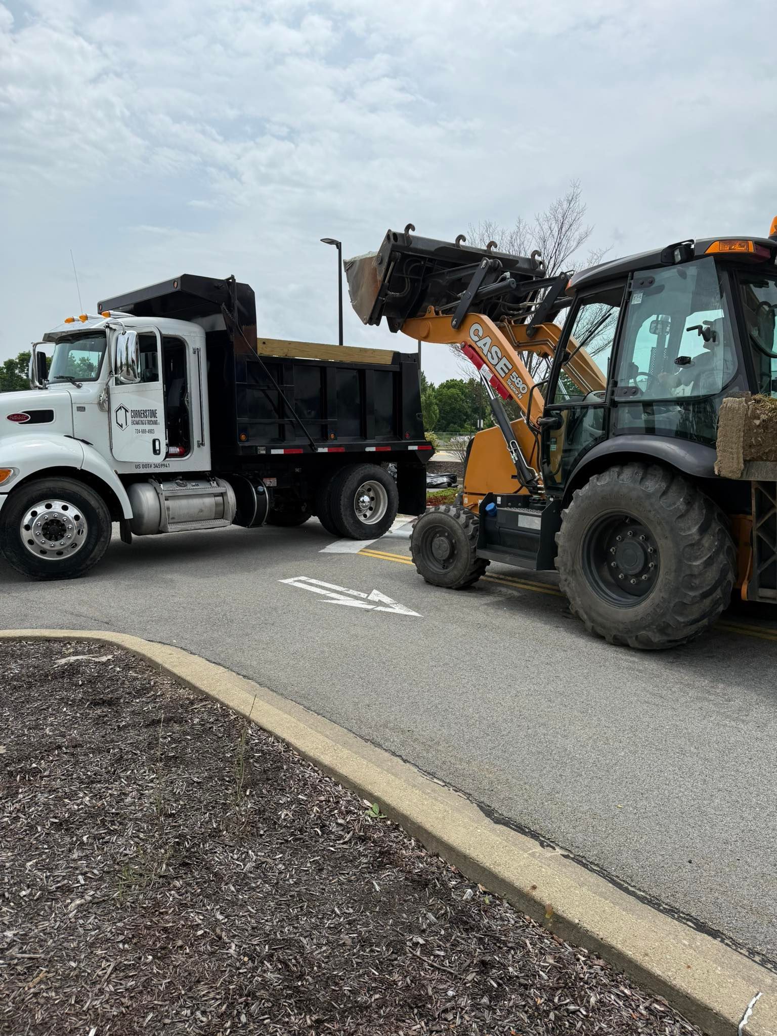 A backhoe loader scoops debris into the bed of a white dump truck on a paved lot. Cloudy sky.