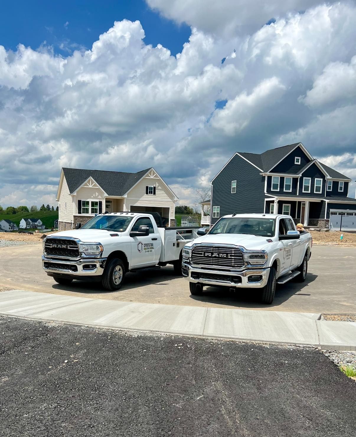 Two white trucks parked in front of new houses on a sunny day. A pile of gravel sits in the foreground.