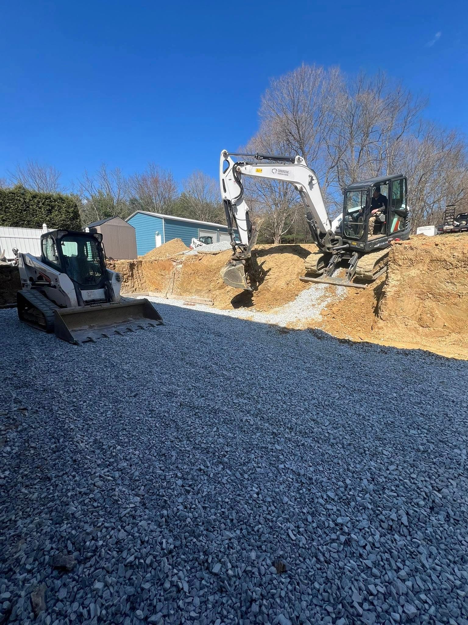 Two construction vehicles, a Bobcat and an excavator, dig in a gravel-covered work site on a sunny day.