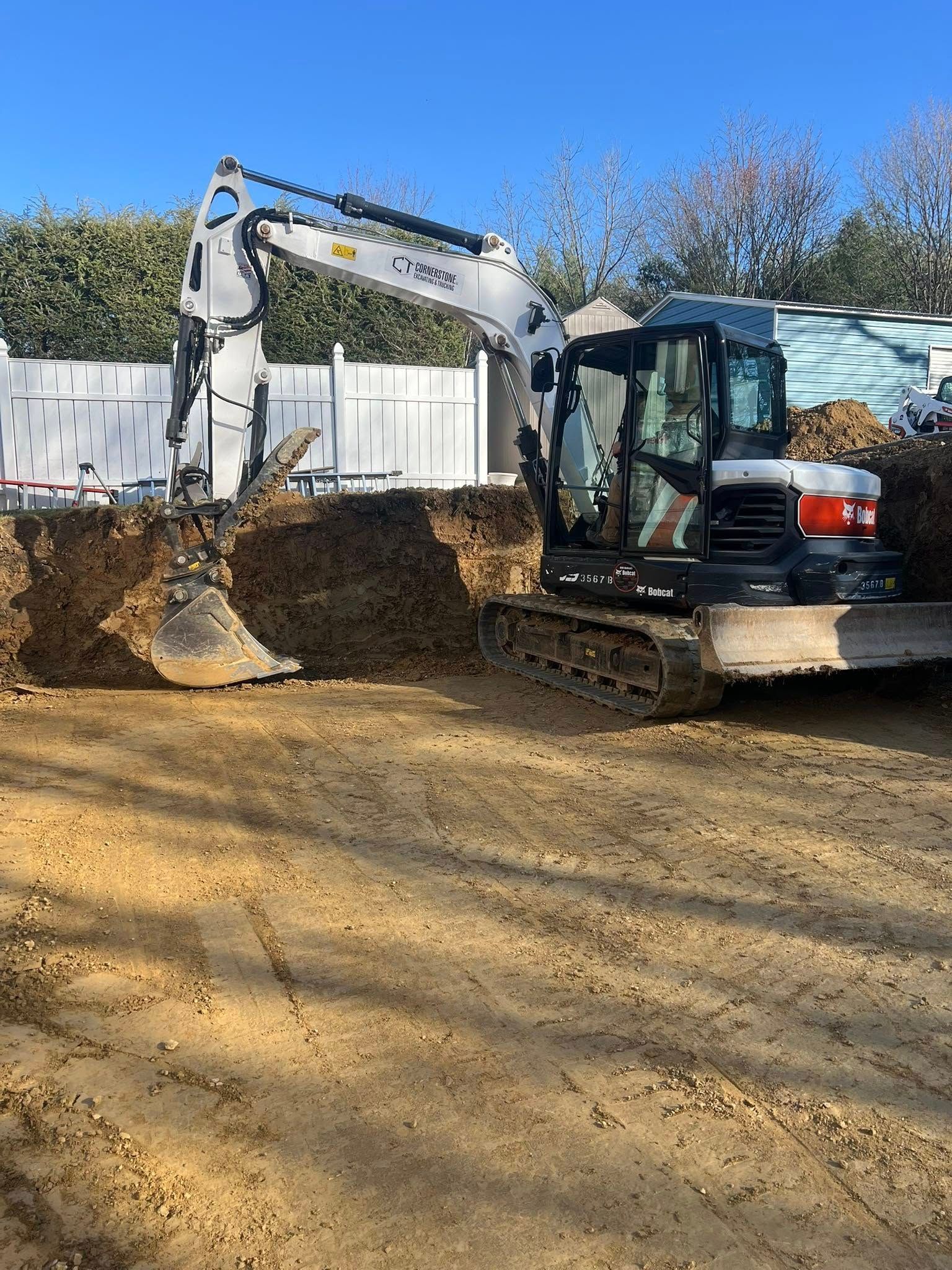 An excavator digs into a brown earth surface next to a white fence under a blue sky.