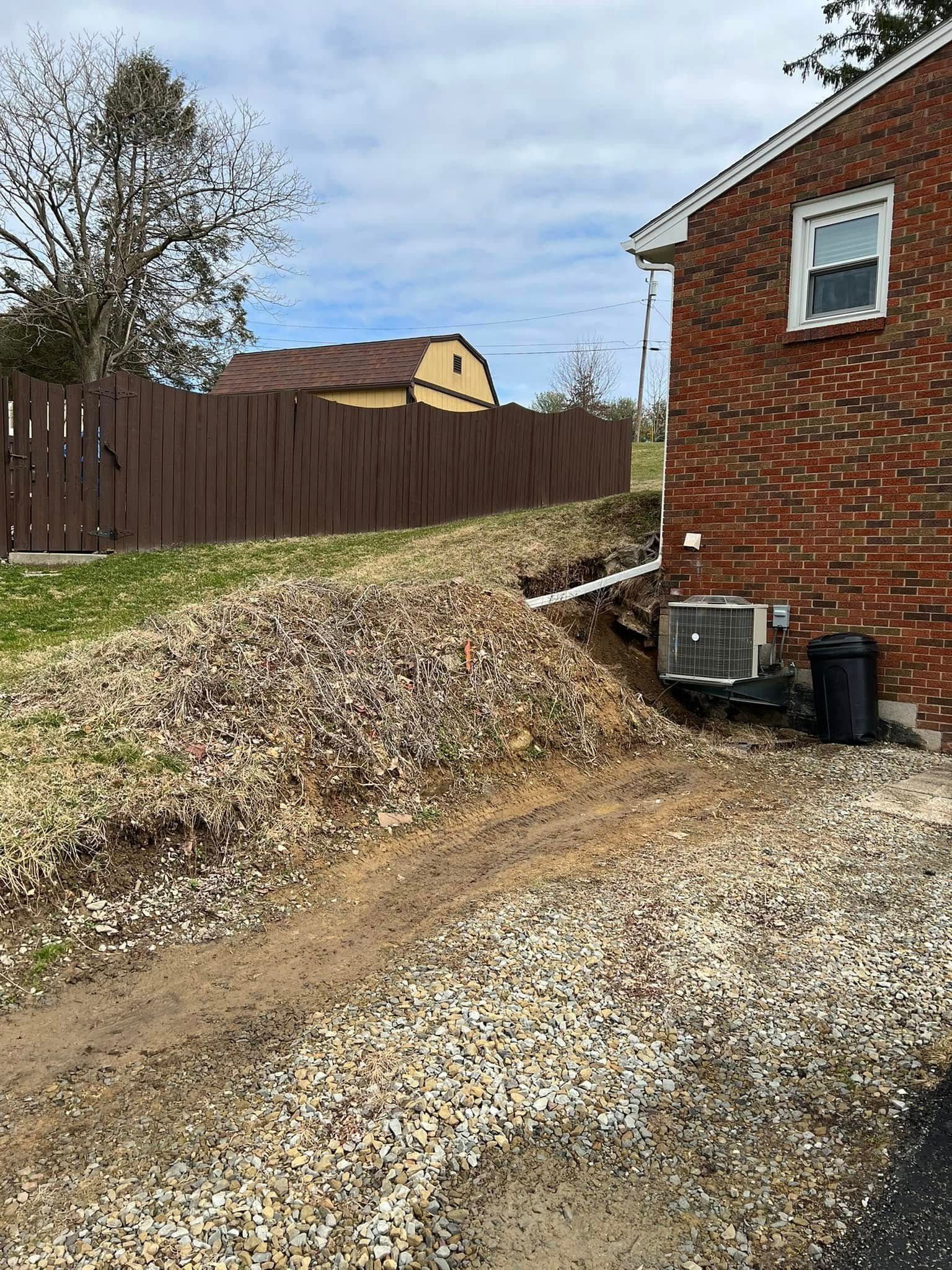 A residential scene with a brick house and a gravel path. A brown fence and barn are in the background under a cloudy sky.