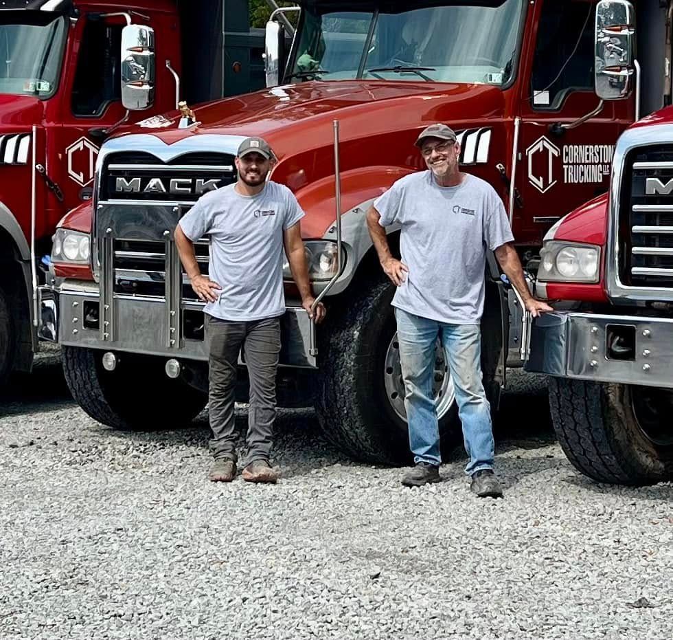Two men stand in front of three red Mack trucks. They are wearing grey t-shirts, smiling and posing for a photo.