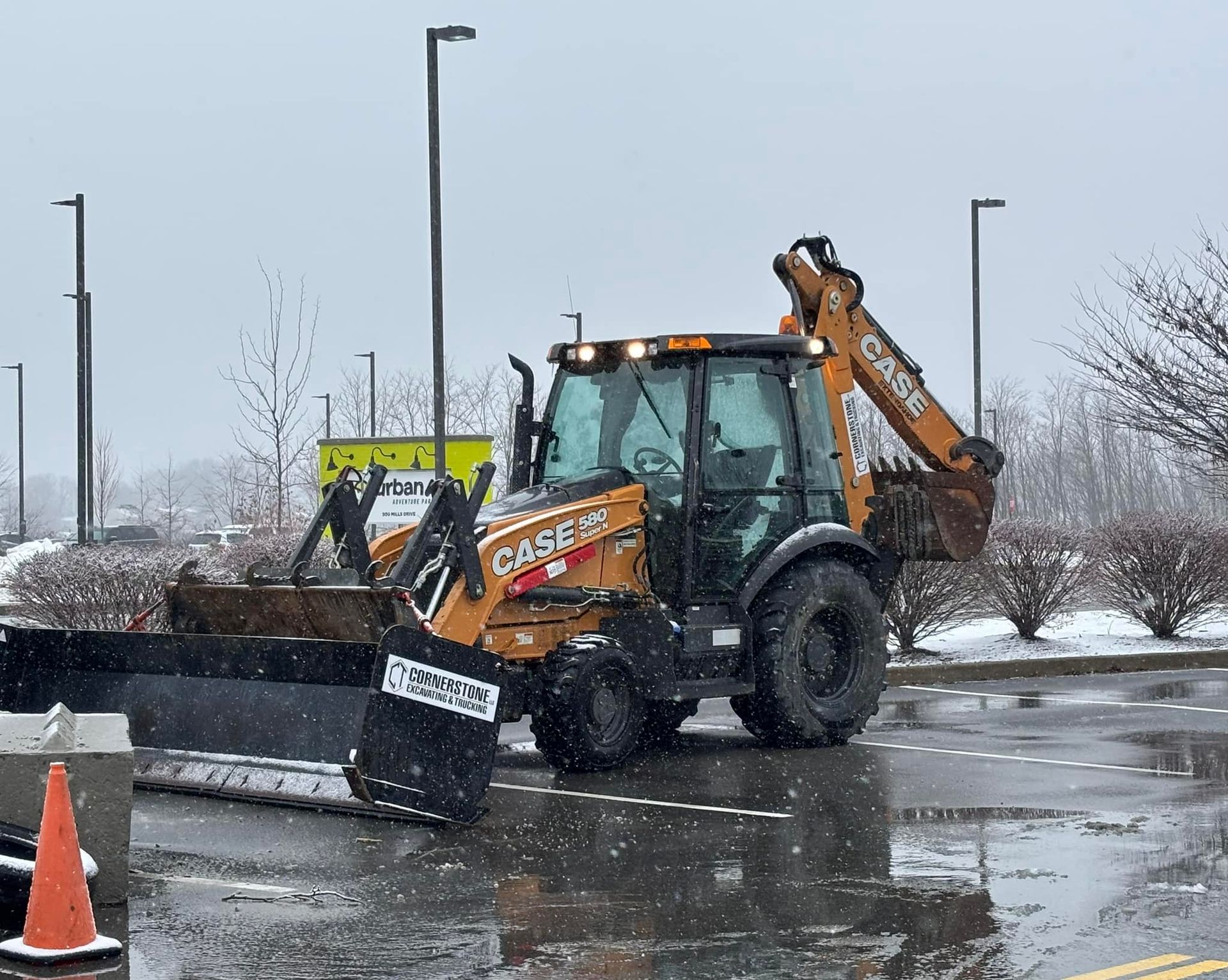 Orange Case backhoe with a snowplow blade clearing a wet parking lot during a snowfall.