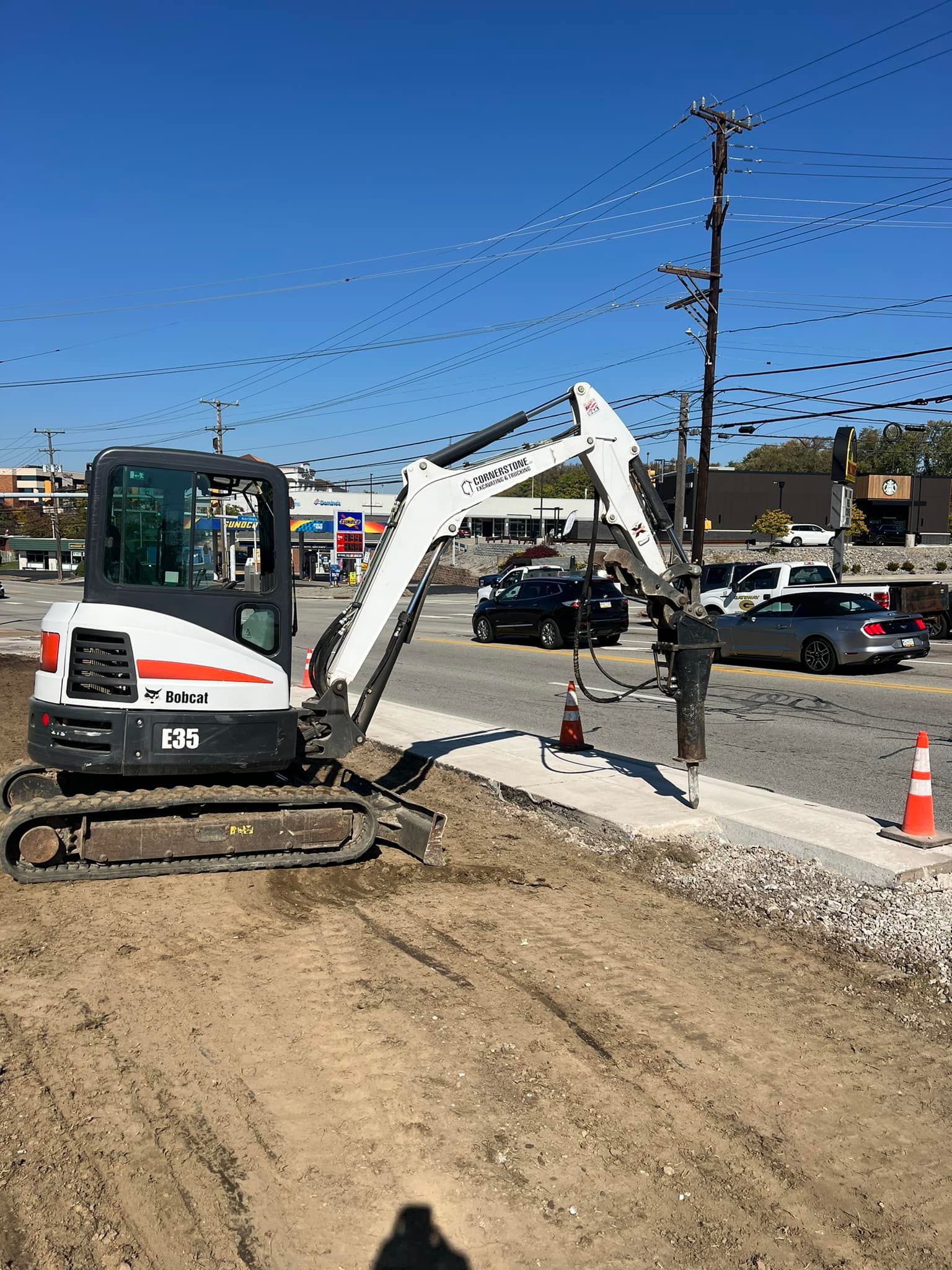 A Bobcat excavator works next to a road, likely for construction. It is a sunny day.