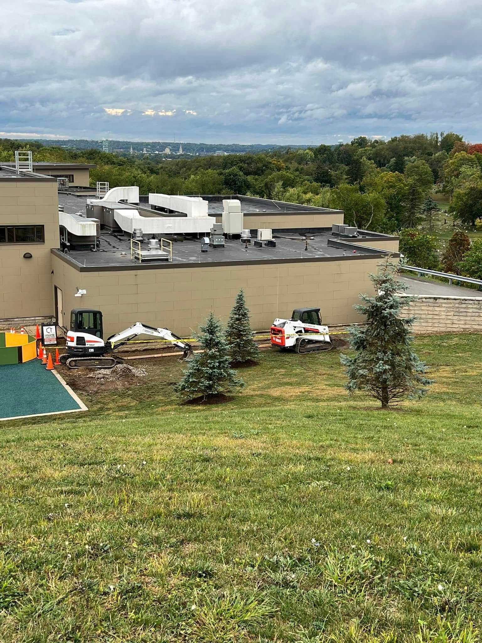 A construction site with heavy machinery next to a light brown building on a grassy hill under a cloudy sky.