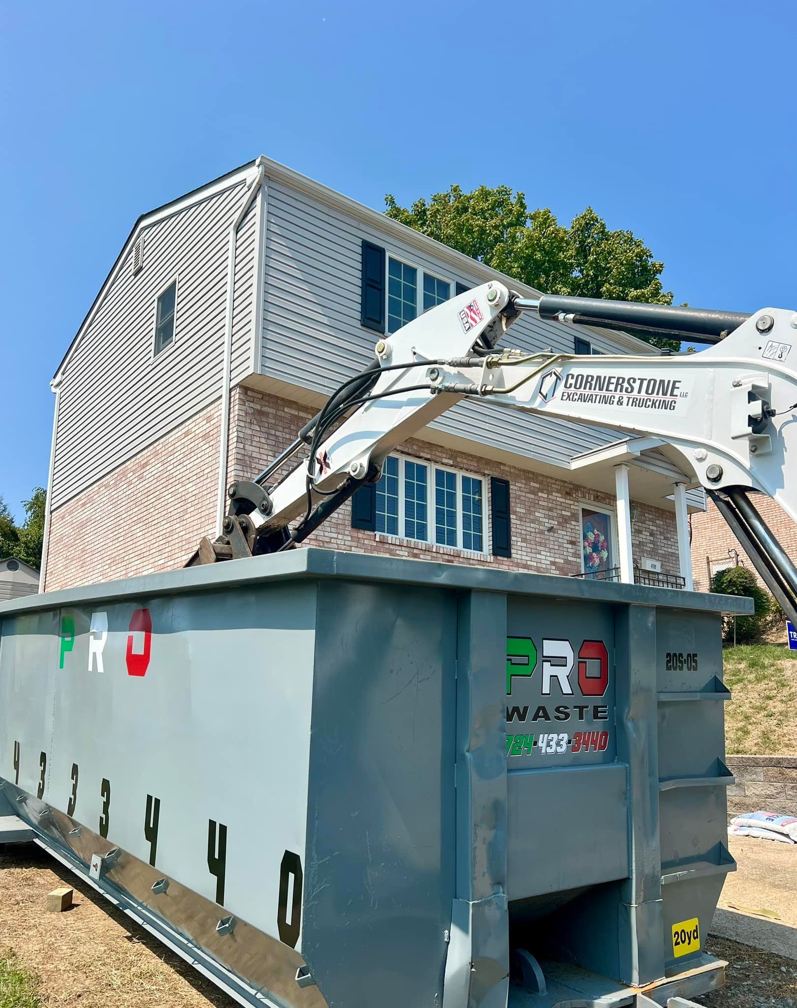 A house being demolished with a mechanical arm, with a large dumpster in the foreground. The scene is outdoors on a sunny day.