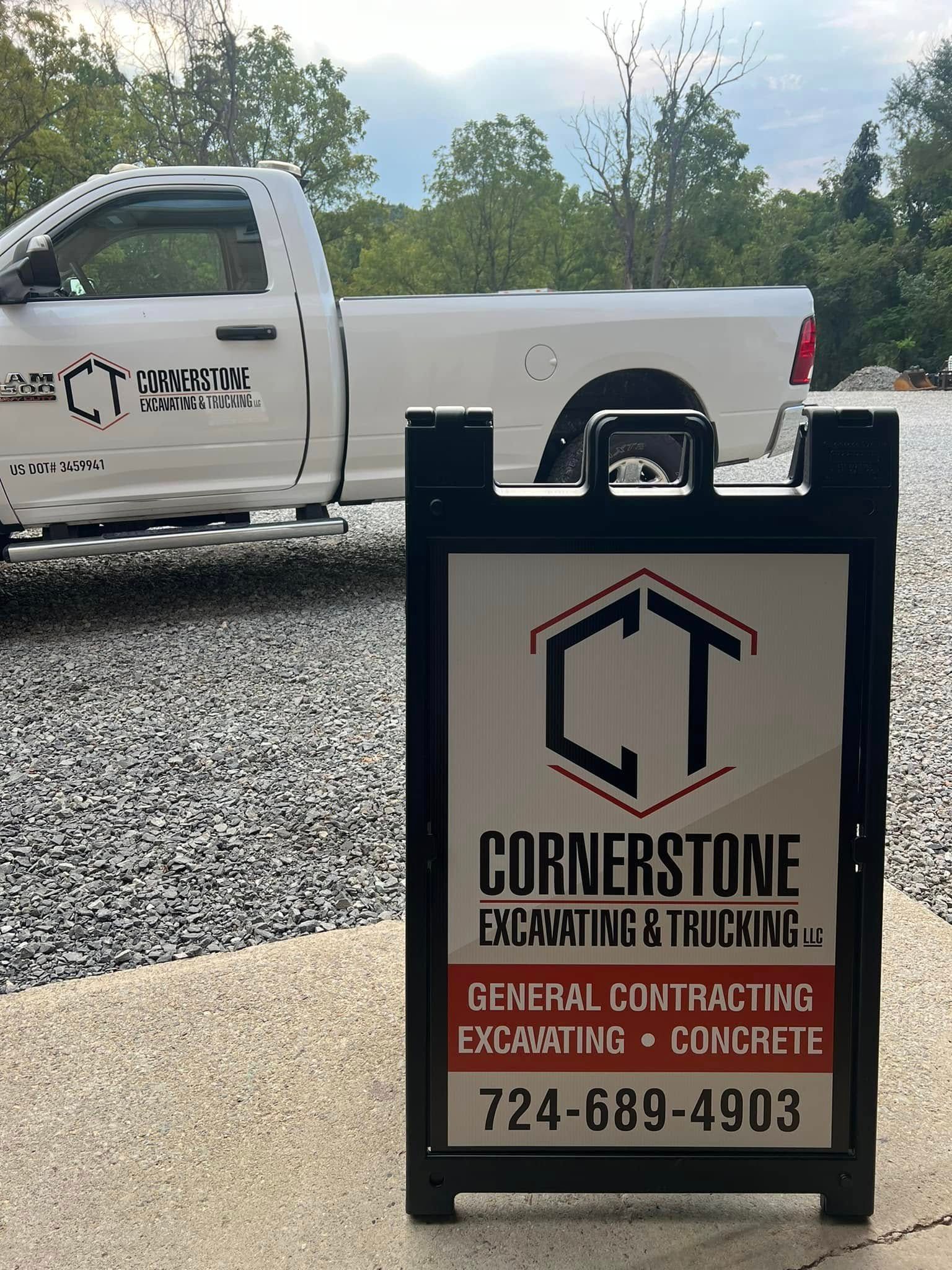 A black A-frame sign for Cornerstone Excavating & Trucking, in front of a white pickup truck on gravel. The sign features a logo and contact information.