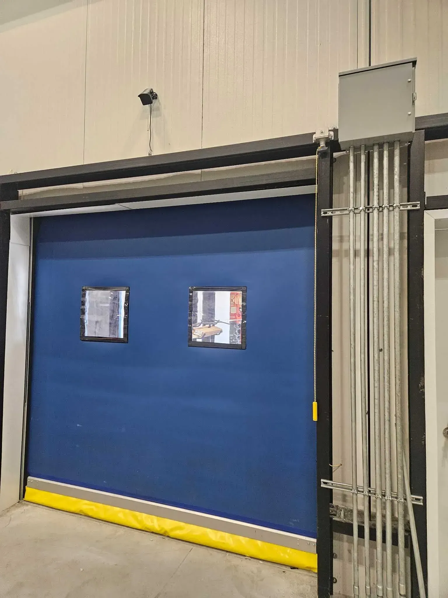 Blue roll-up door with yellow bottom and two windows. Steel tracks and control box on the side, set indoors.