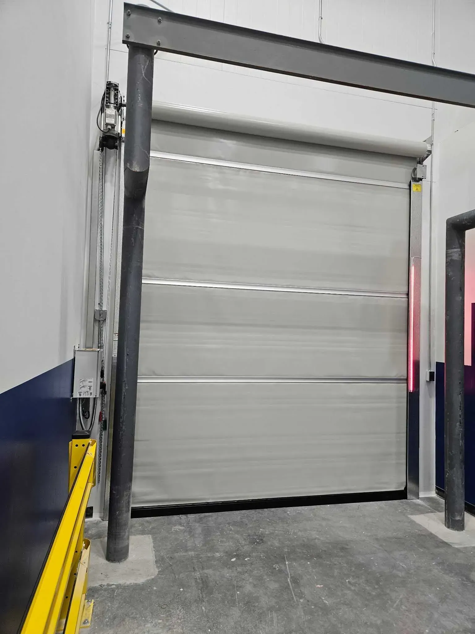 Gray industrial roll-up door in an interior warehouse setting, with metal frame and yellow safety bar.