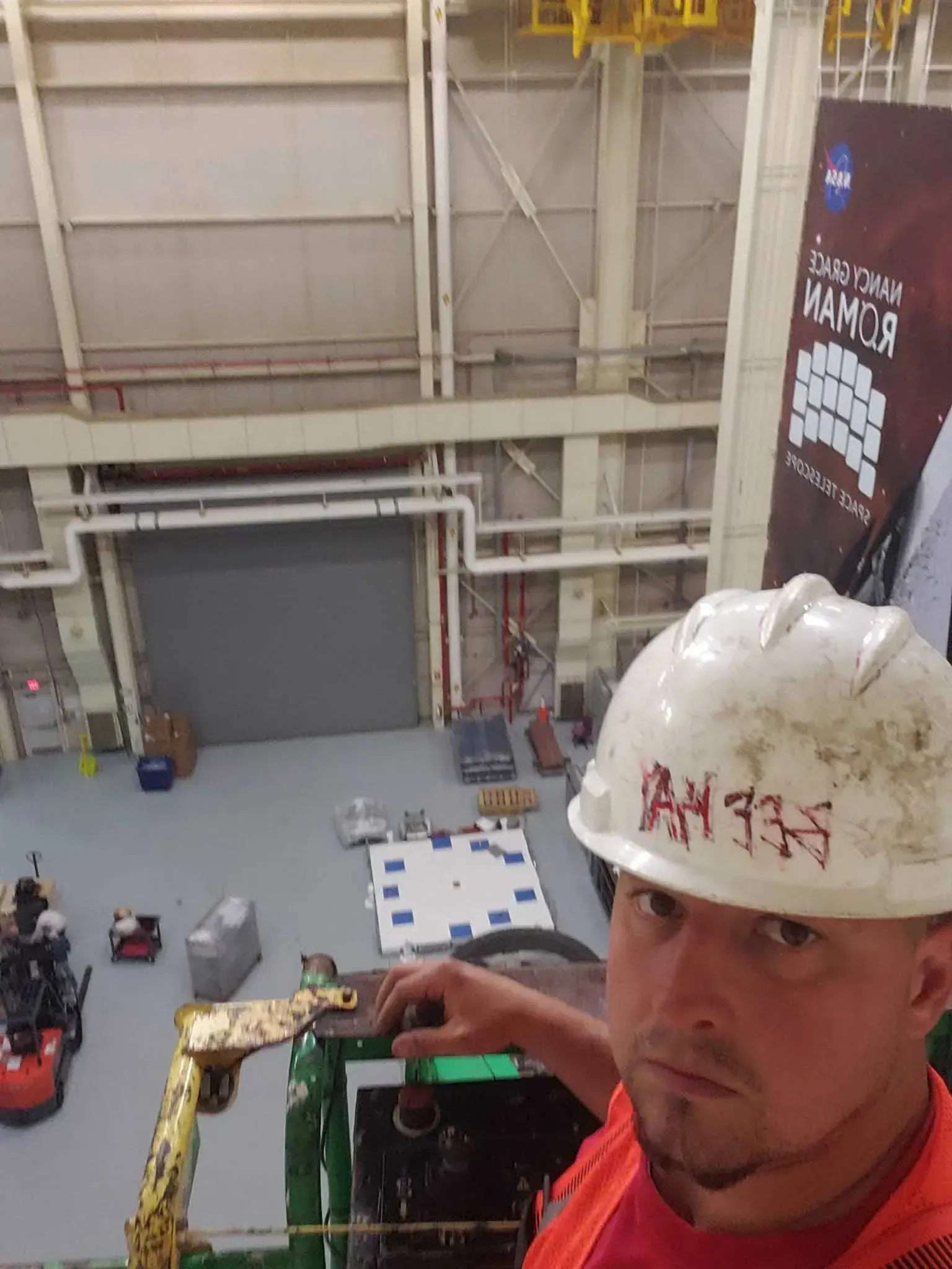 Man in hard hat, orange vest looks down from lift in large hangar. Equipment on floor, NASA banner.