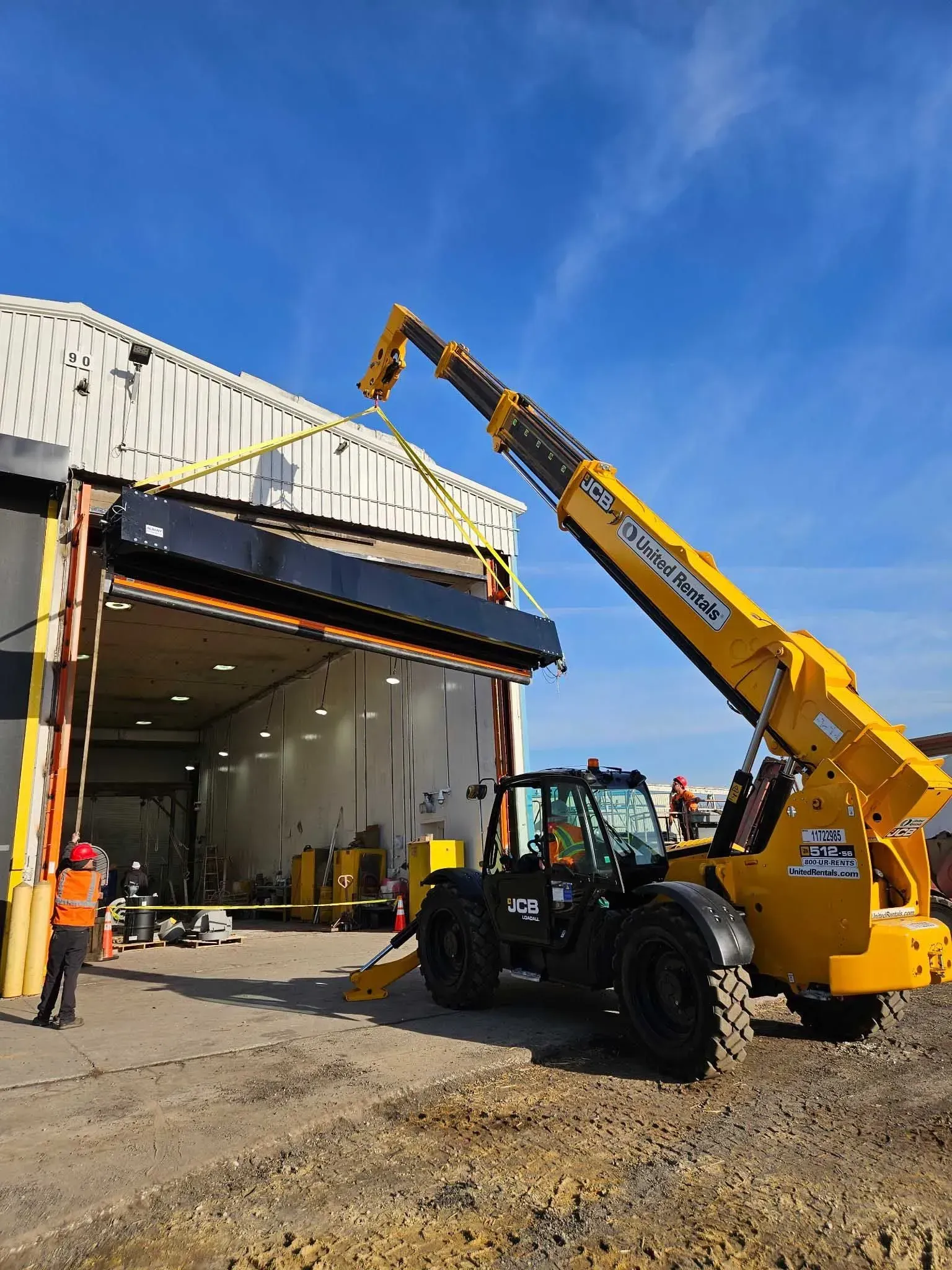 Yellow telehandler lifting a black structure into a warehouse opening on a sunny day.