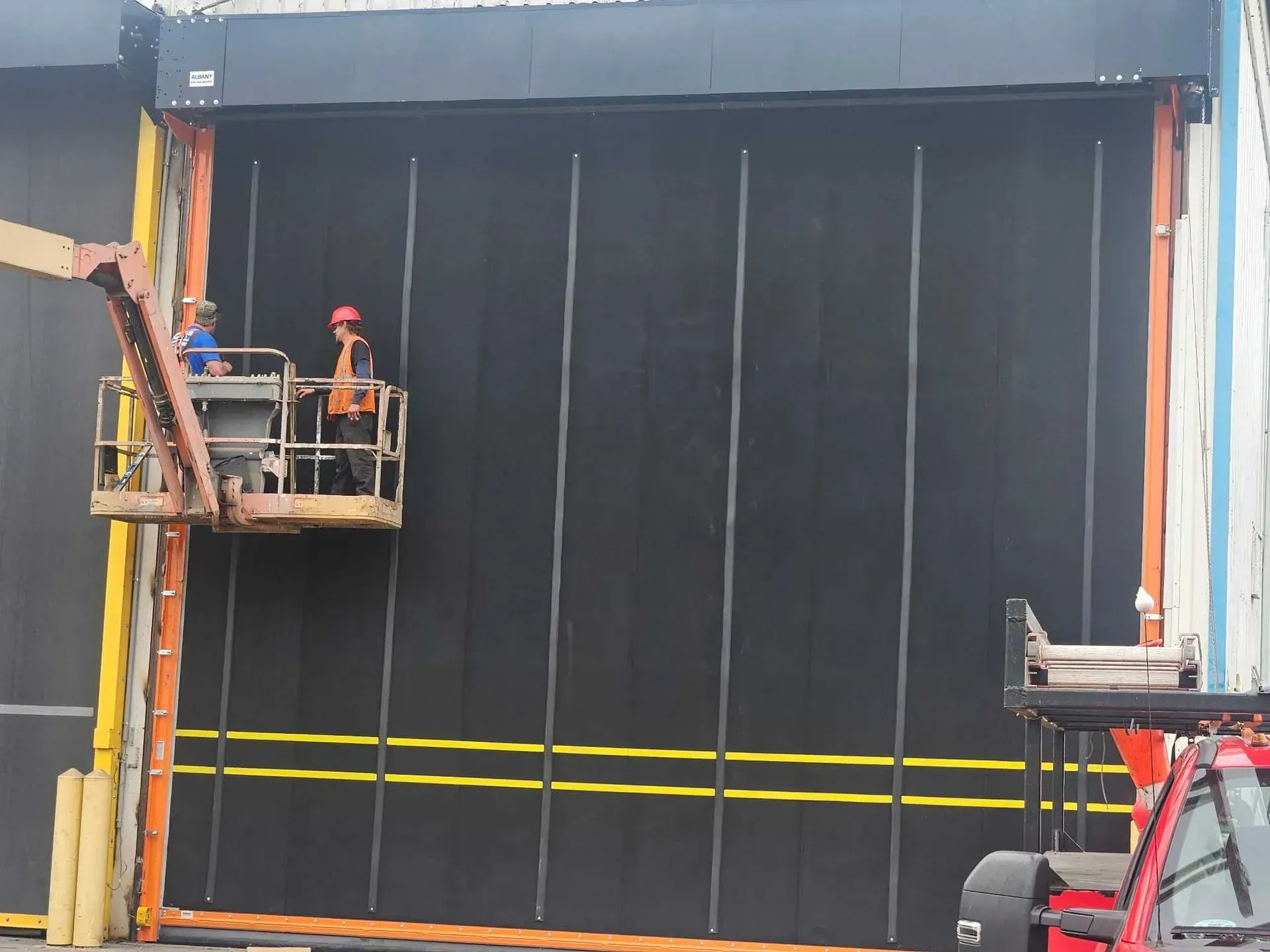 Two workers in a lift installing a large, black, industrial door on a building.
