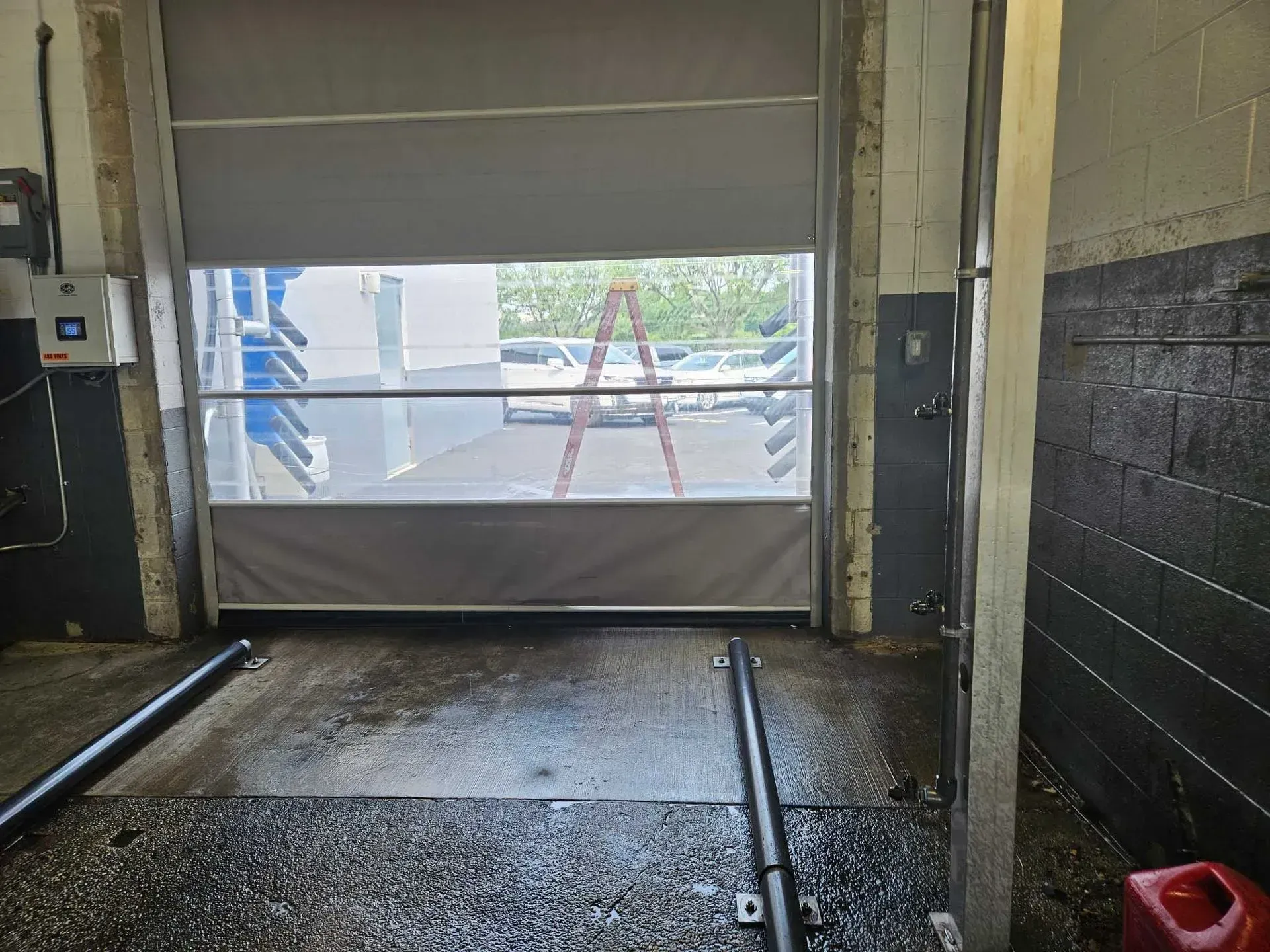 Garage bay with grey roll-up door partially open, showing a ladder outside. Wet floor, concrete walls.
