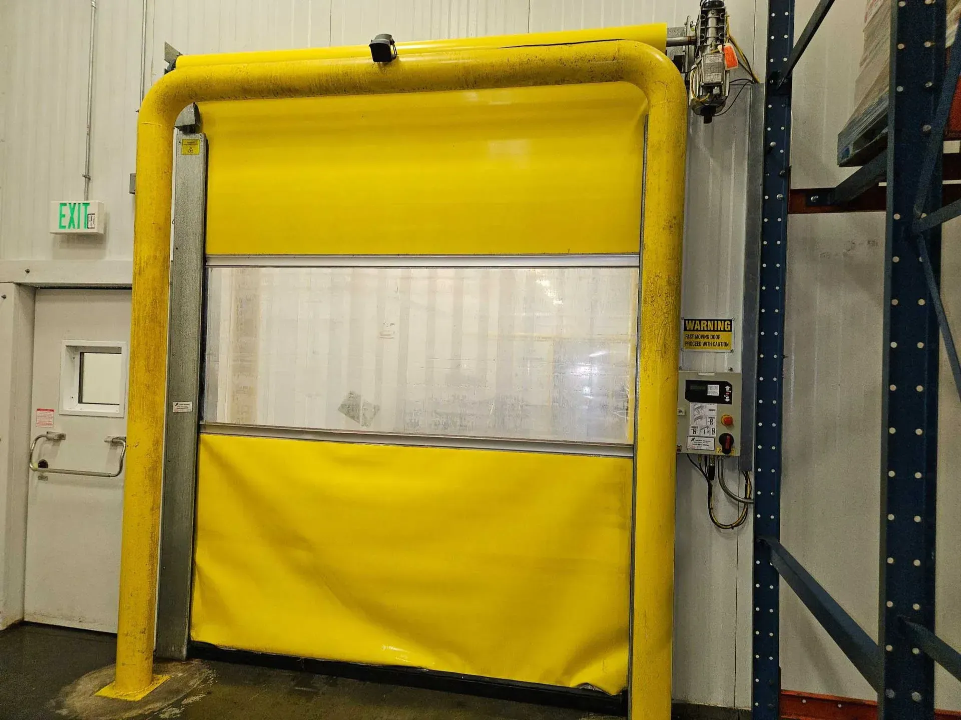 Yellow high-speed roll-up door in a warehouse setting, flanked by yellow safety bars.