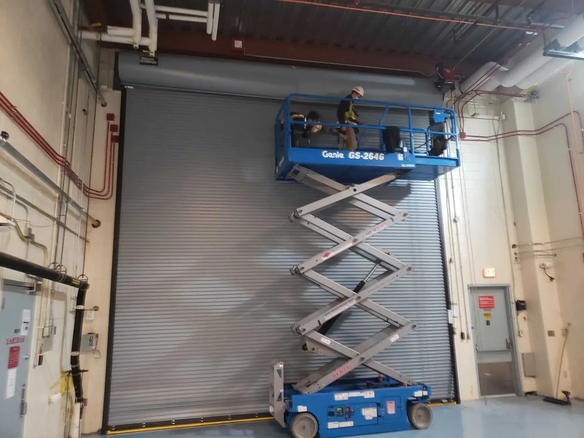 A worker on a blue scissor lift is working on a large gray overhead door inside a building.