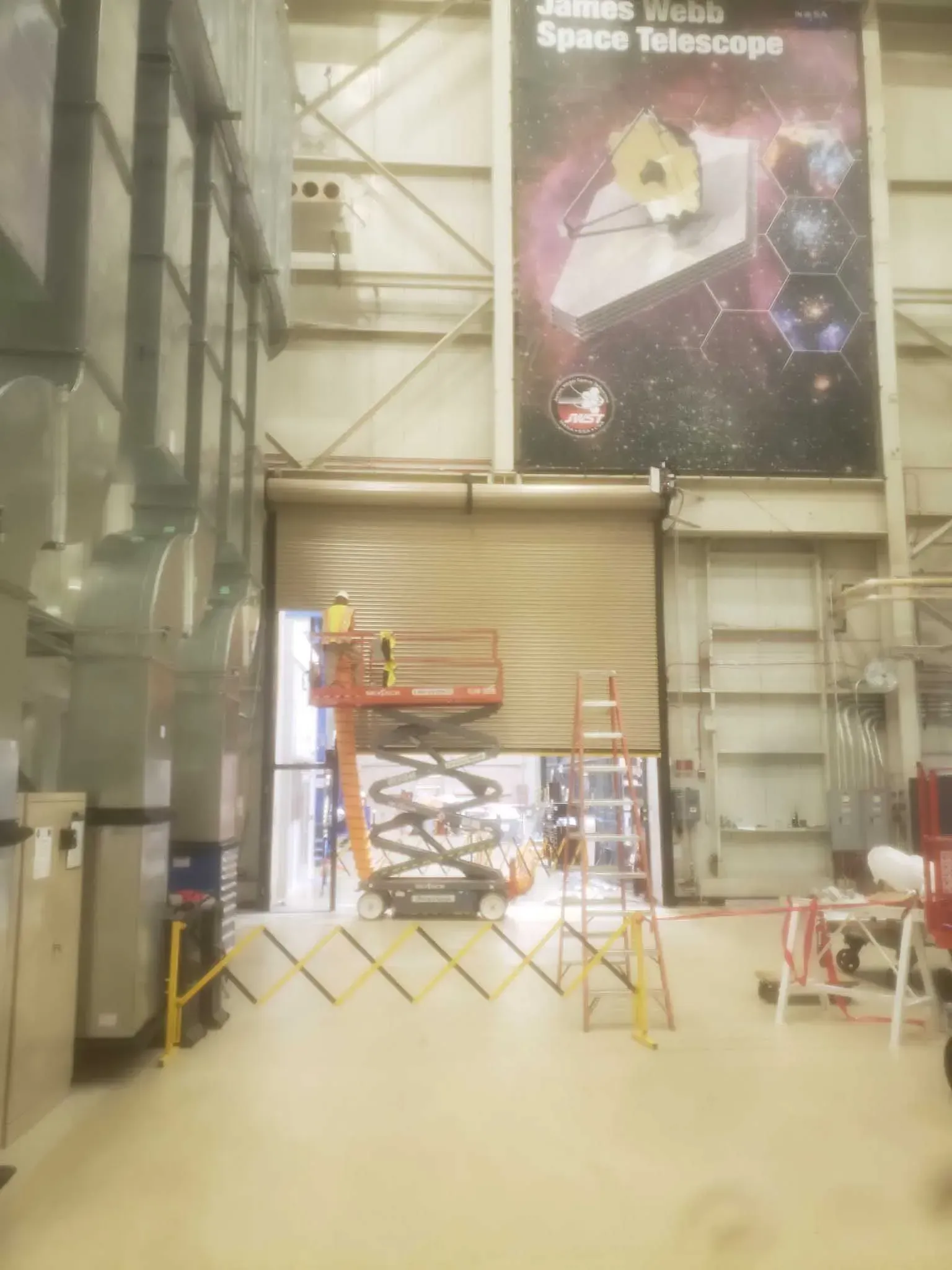 Inside a warehouse, workers on a lift and a ladder near a partially opened garage door, with a James Webb Space Telescope banner.