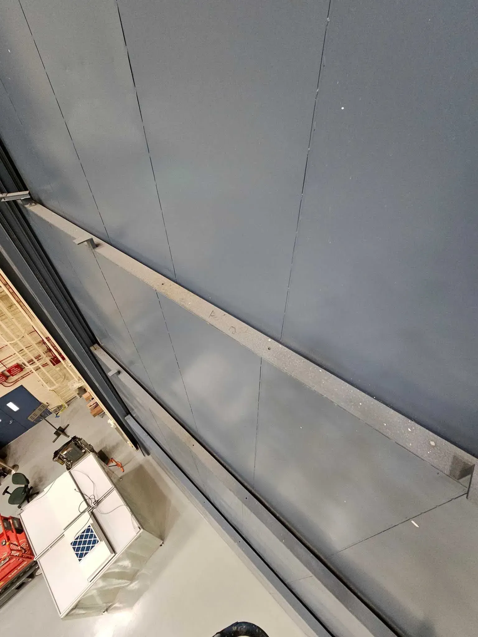 Looking up at a gray wall, with angled silver metal support structure, with a few objects and other wall in the background.