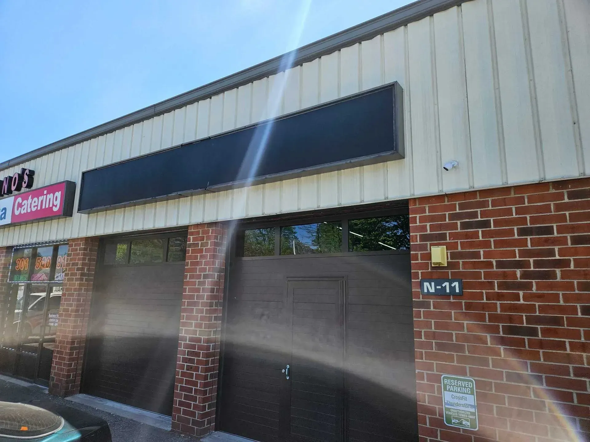 Storefront with black sign, brown brick, and garage doors. Bright sun.