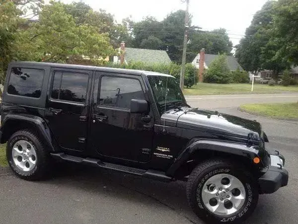 Black Jeep Wrangler parked on a driveway, trees and houses in background.