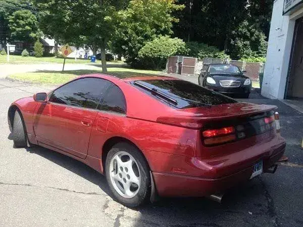 Red Nissan 300ZX coupe parked on a paved surface, tinted windows, in front of a garage and another car.