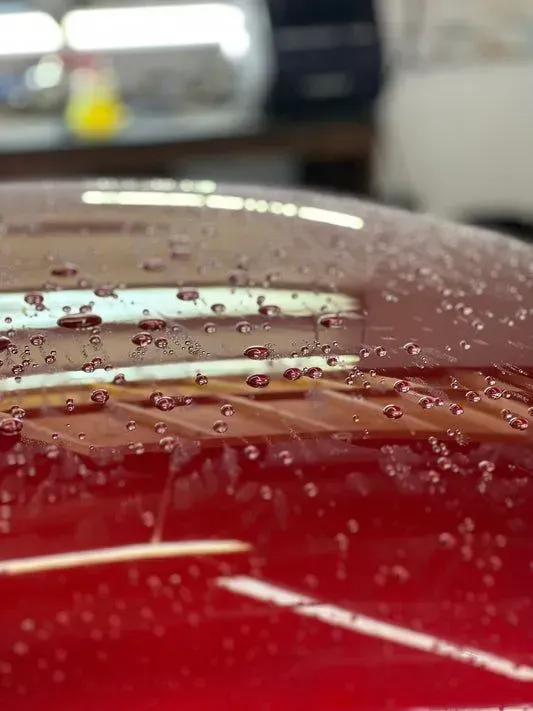 Close-up of a red car roof with water droplets. The droplets are small and round, reflecting the overhead lights.