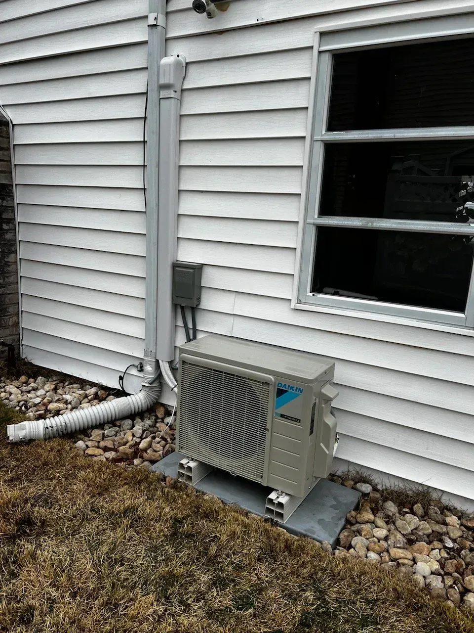 Outdoor HVAC unit beside a white house wall, with a window, downspout, and patchy grass below.