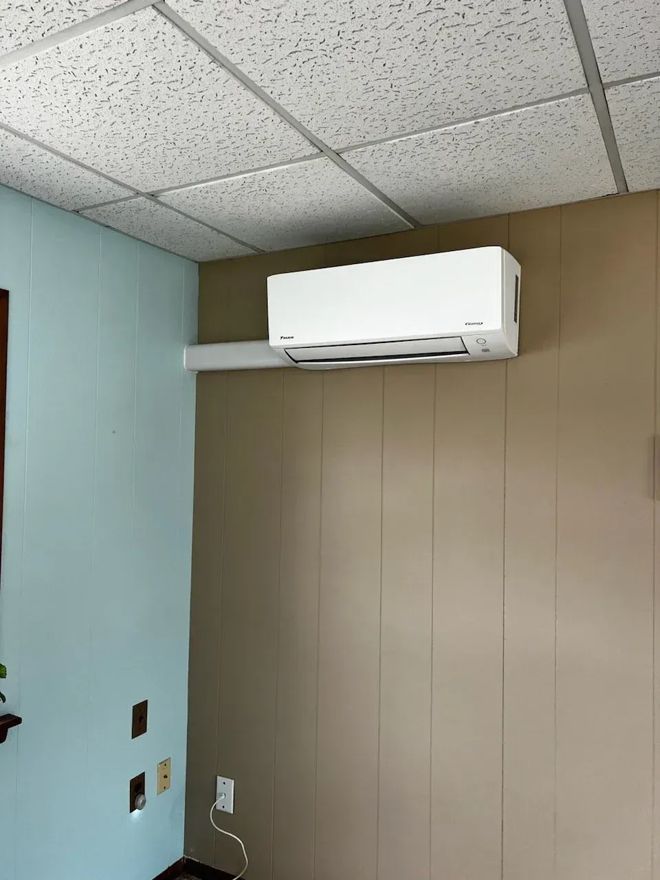 White wall-mounted air conditioner on a beige wall in a small room with drop ceiling tiles.