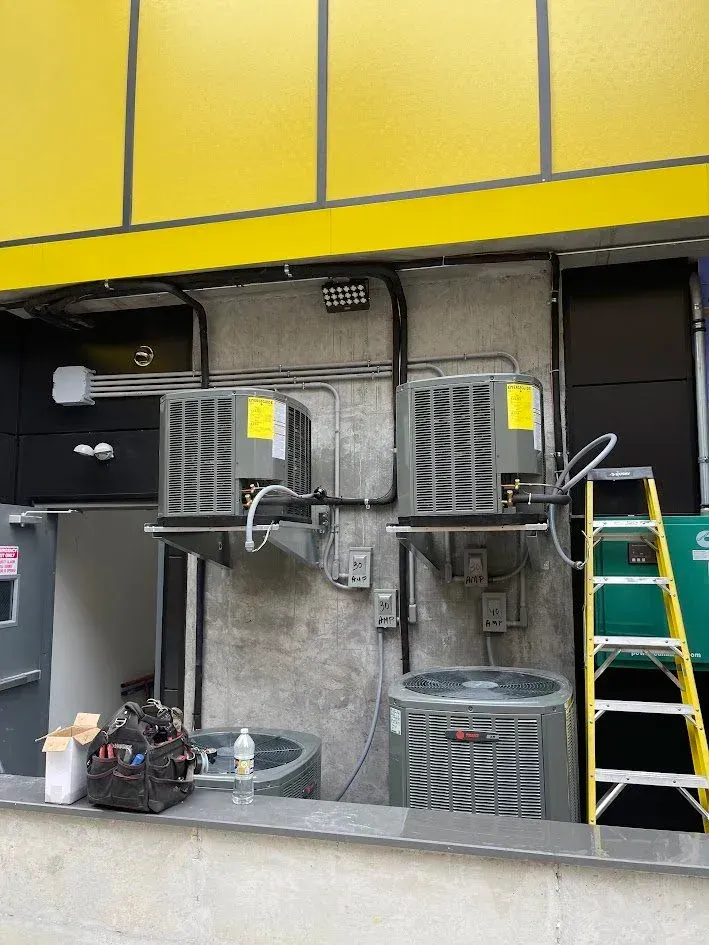 Outdoor wall with two large gray HVAC units, a yellow building panel above, and a ladder at right.