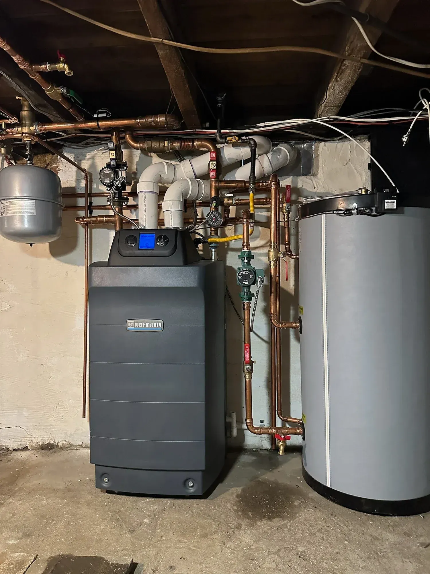 Basement HVAC and water heater setup with dark furnace, gray tank, and exposed pipes under low ceiling