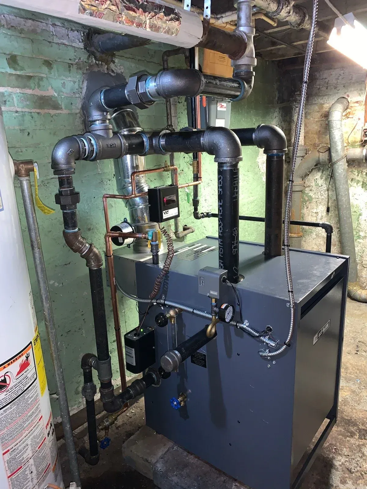 Industrial HVAC/boiler unit with black piping in a narrow basement utility room