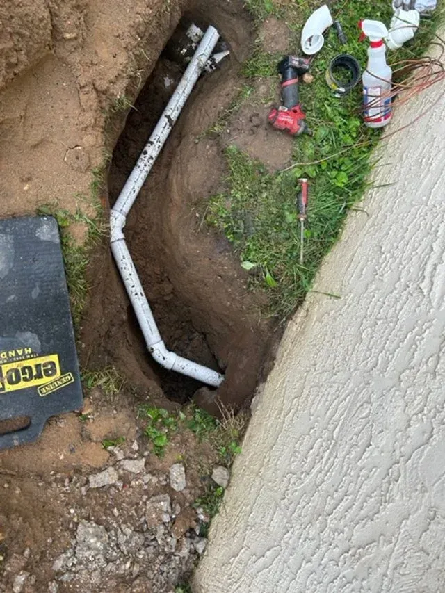 Open trench beside pavement with white PVC pipe, tools, and dirt piled around it