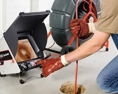 Worker using a drain cleaning machine with a coiled red cable and hose in a room with a floor drain.