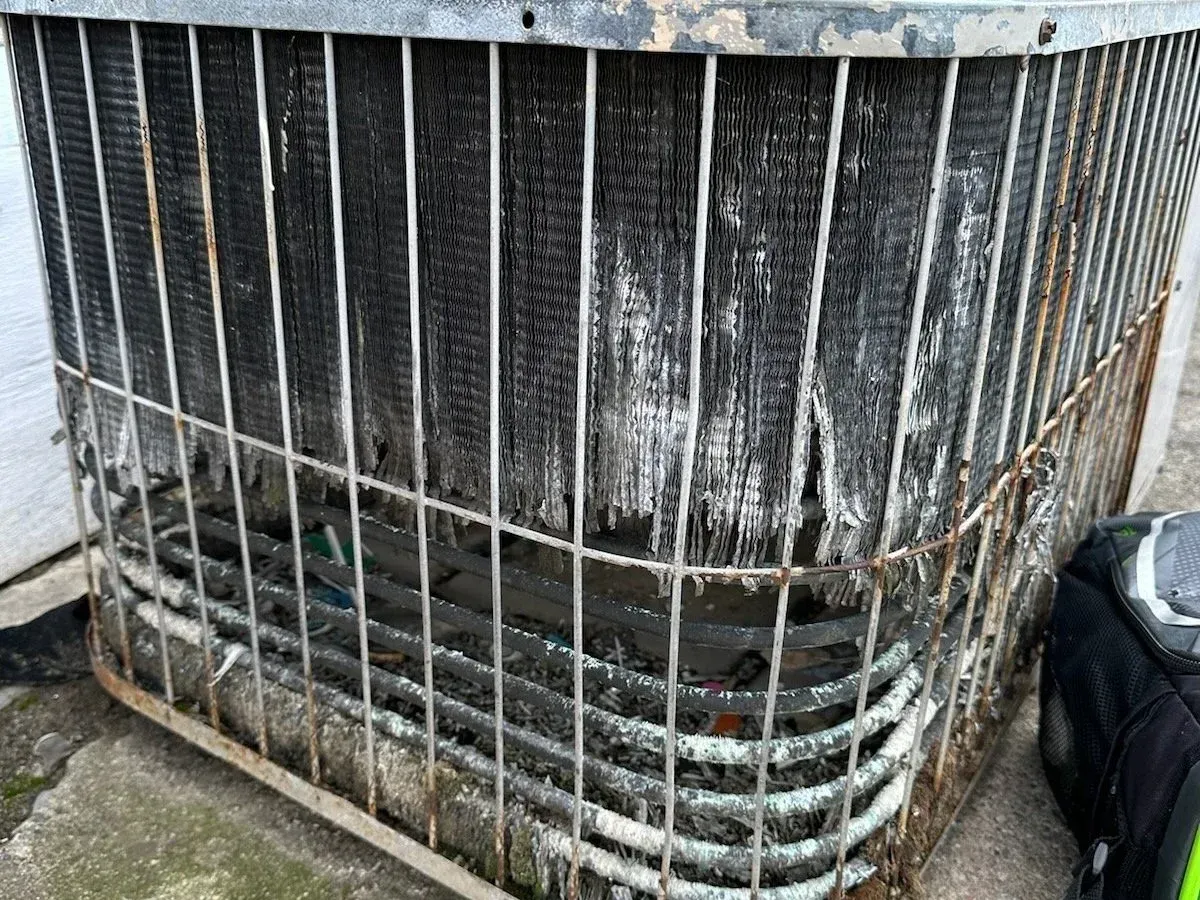 Dirty outdoor air conditioning unit with bent metal grille and debris around the base